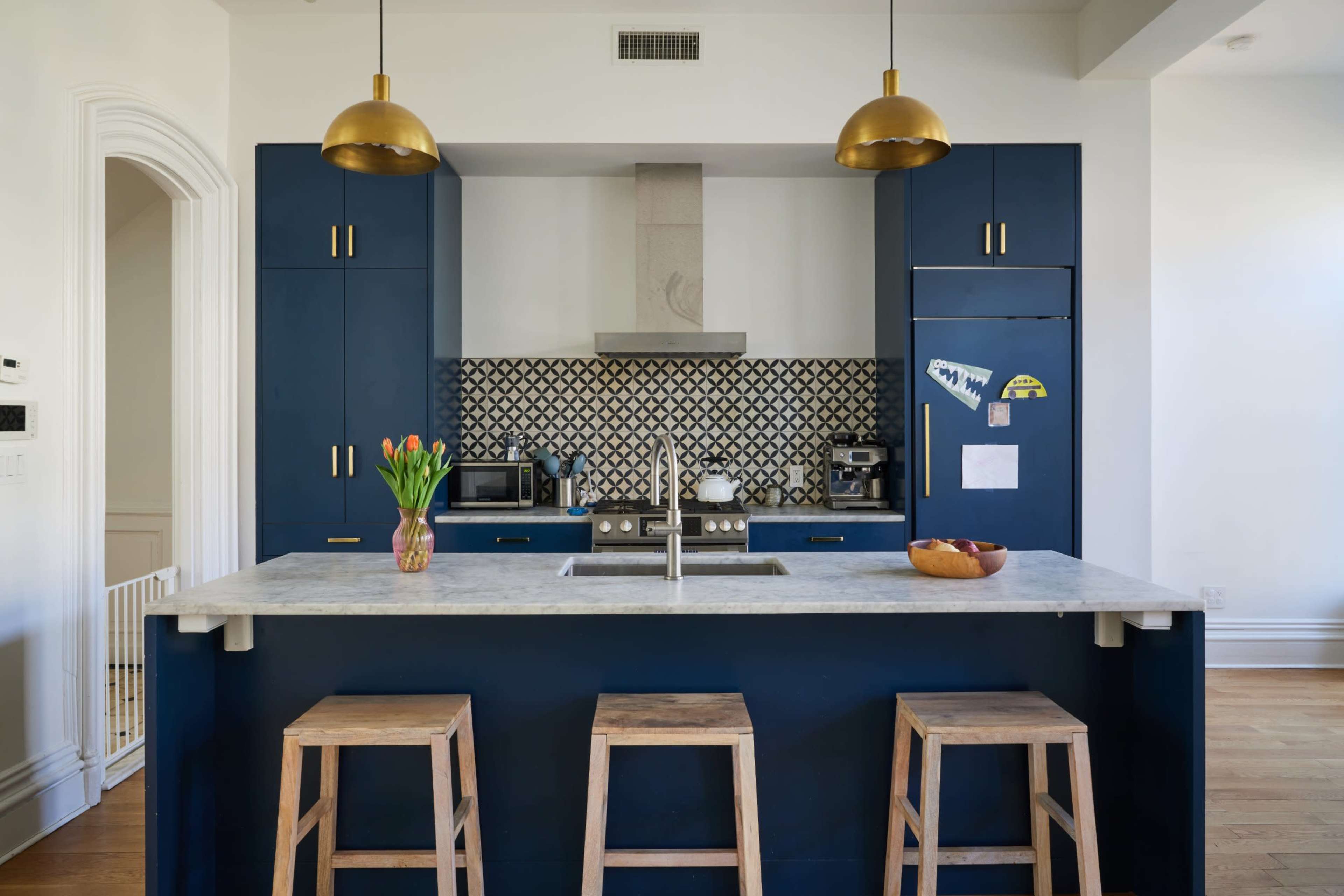 The image shows a modern kitchen featuring blue cabinets, a marble countertop island, and pendant lights above wooden stools.