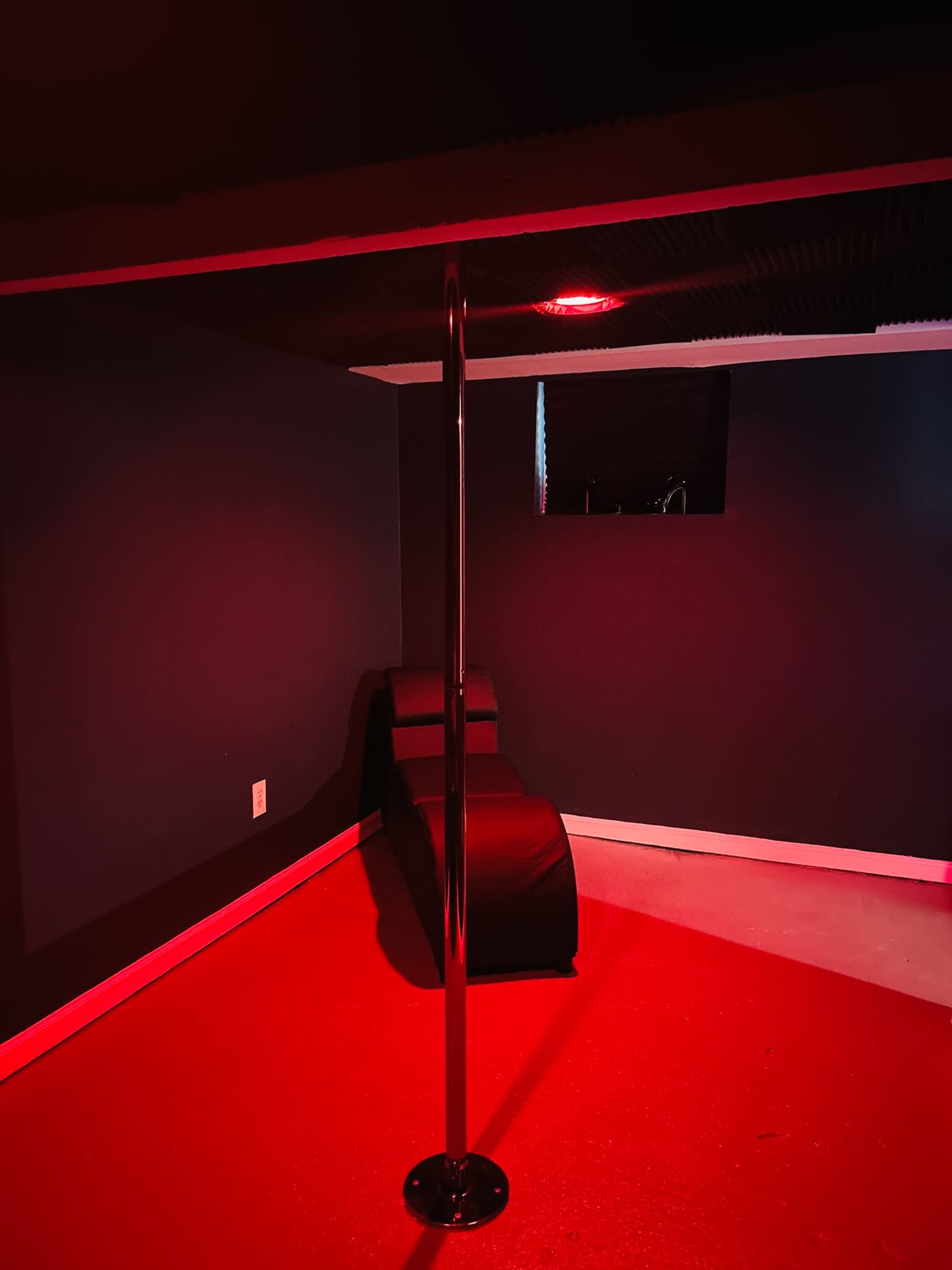 The image shows a dimly lit room with a shiny pole in the center and a black bench on one side, highlighted by red lighting.