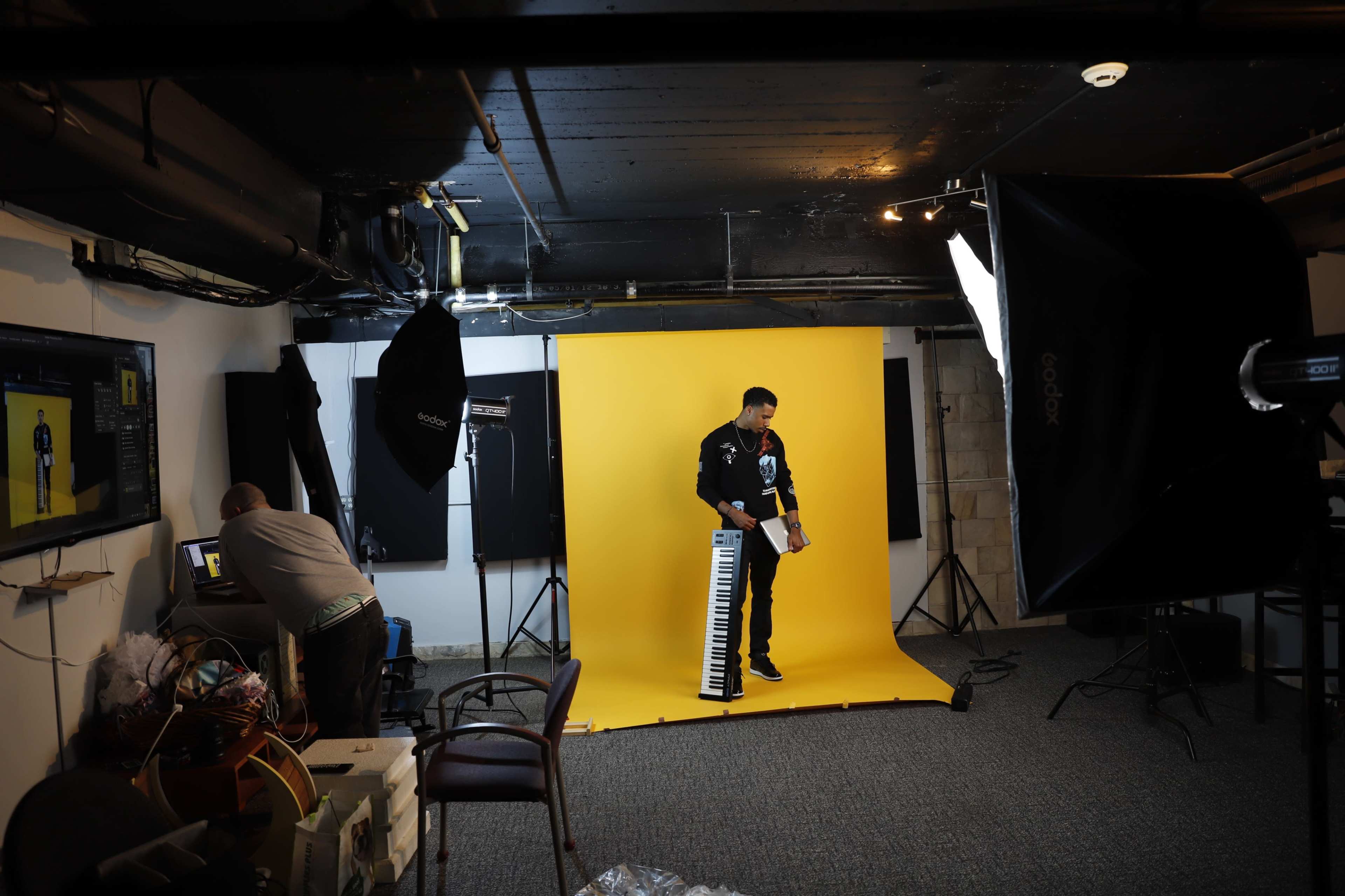 A musician stands with a keyboard in front of a yellow backdrop while a photographer edits images on a computer in a studio setting.