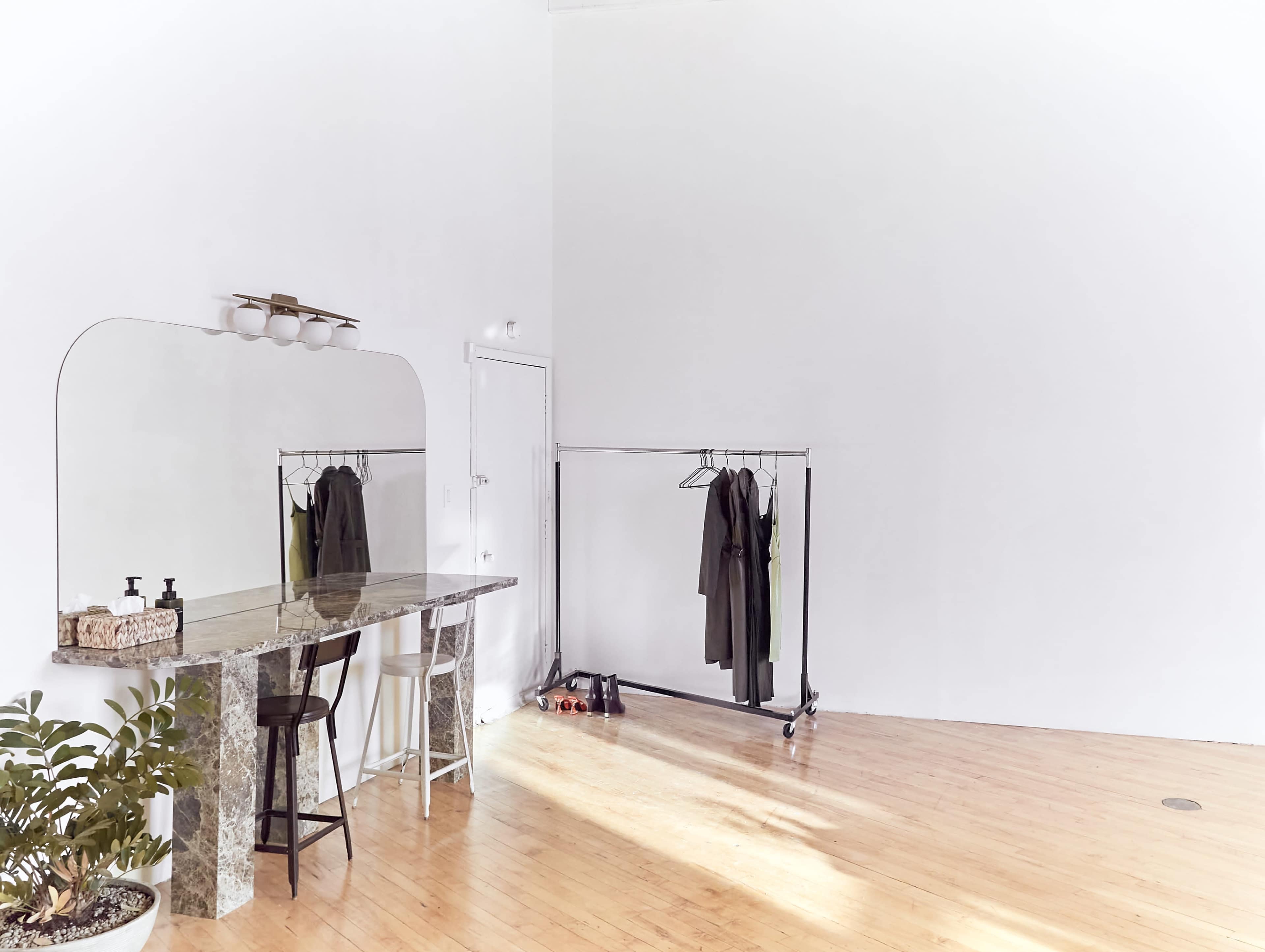 A minimalist dressing area features a mirror, a marble table, a clothing rack with garments, and a small plant in a light-filled room with wooden flooring.