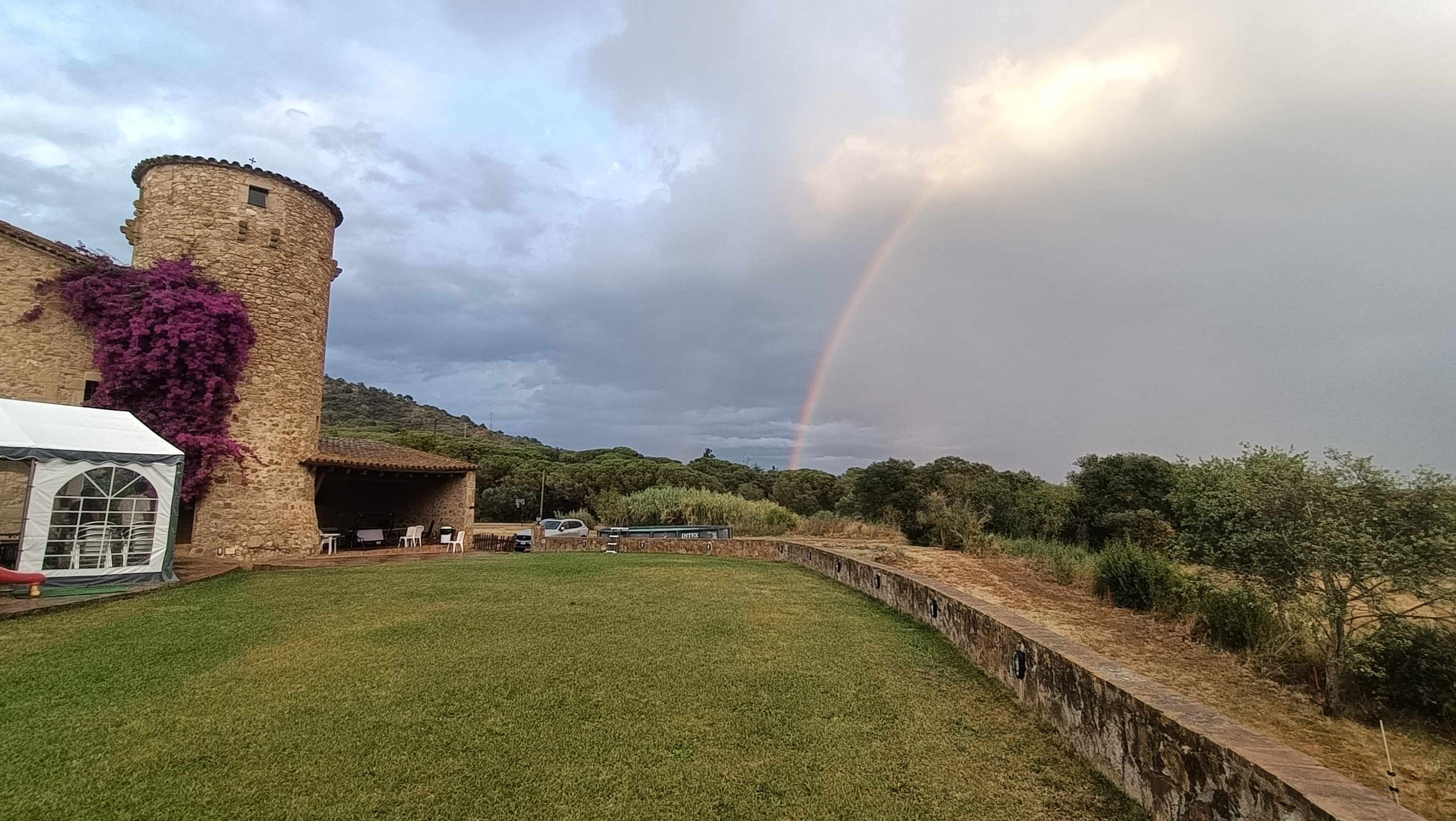 A stone tower covered in purple flowers stands next to a grassy area, with a rainbow visible in the background against a cloudy sky.
