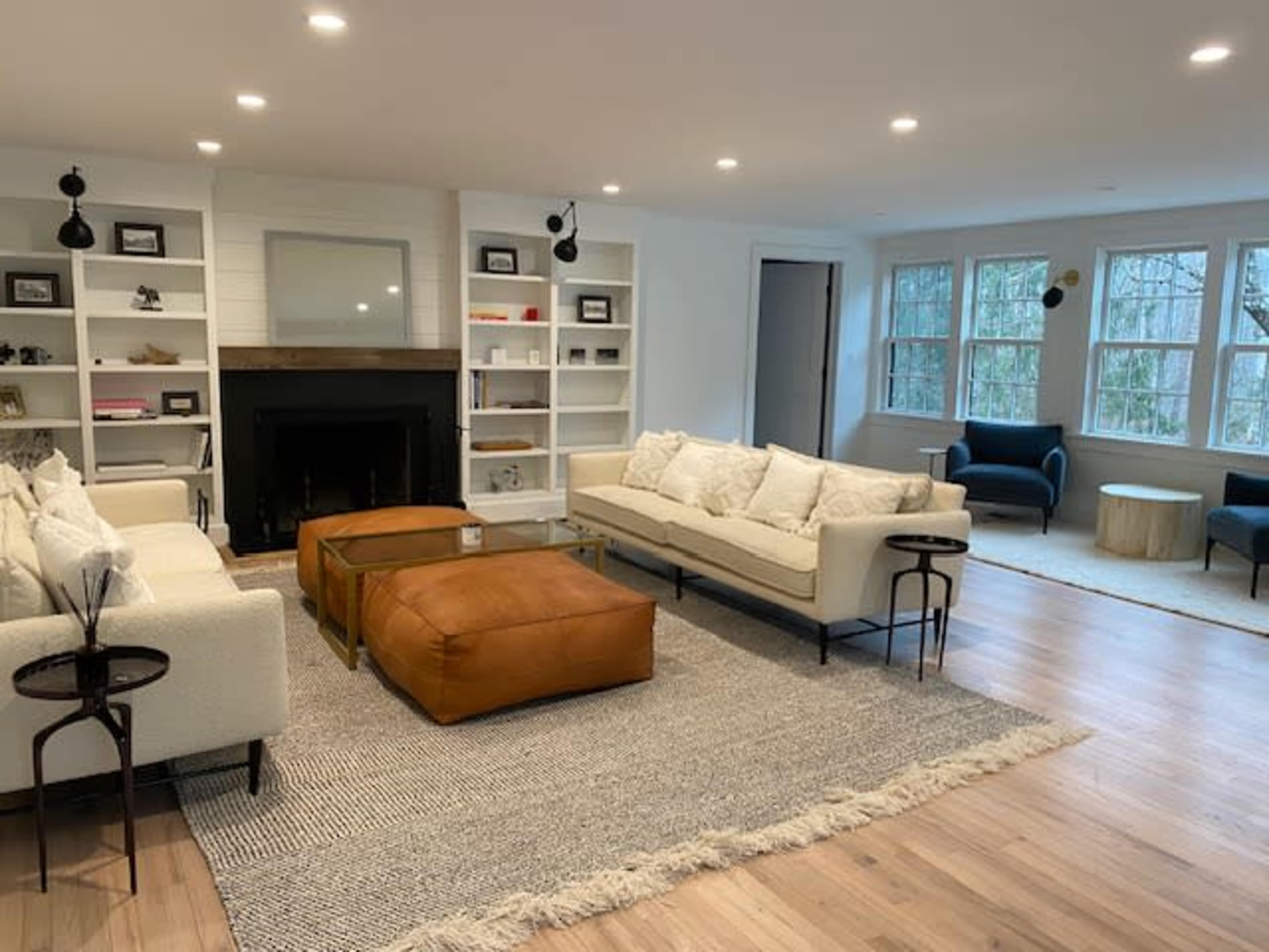 The living room features two sofas facing a coffee table, flanked by bookshelves and a fireplace, with large windows letting in natural light.