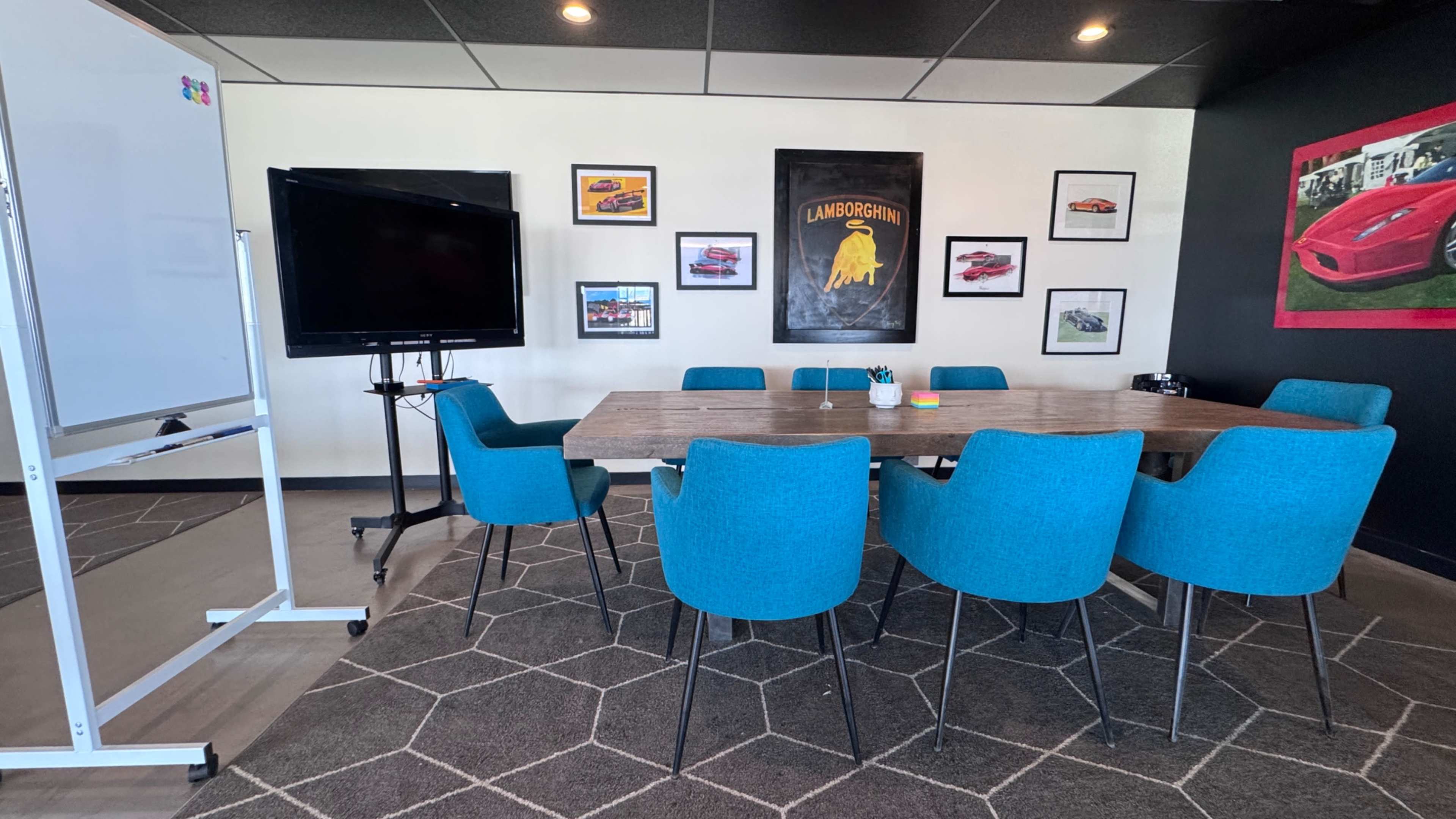 A conference room features a large wooden table surrounded by blue chairs, a television on a stand, and framed car posters on the walls.