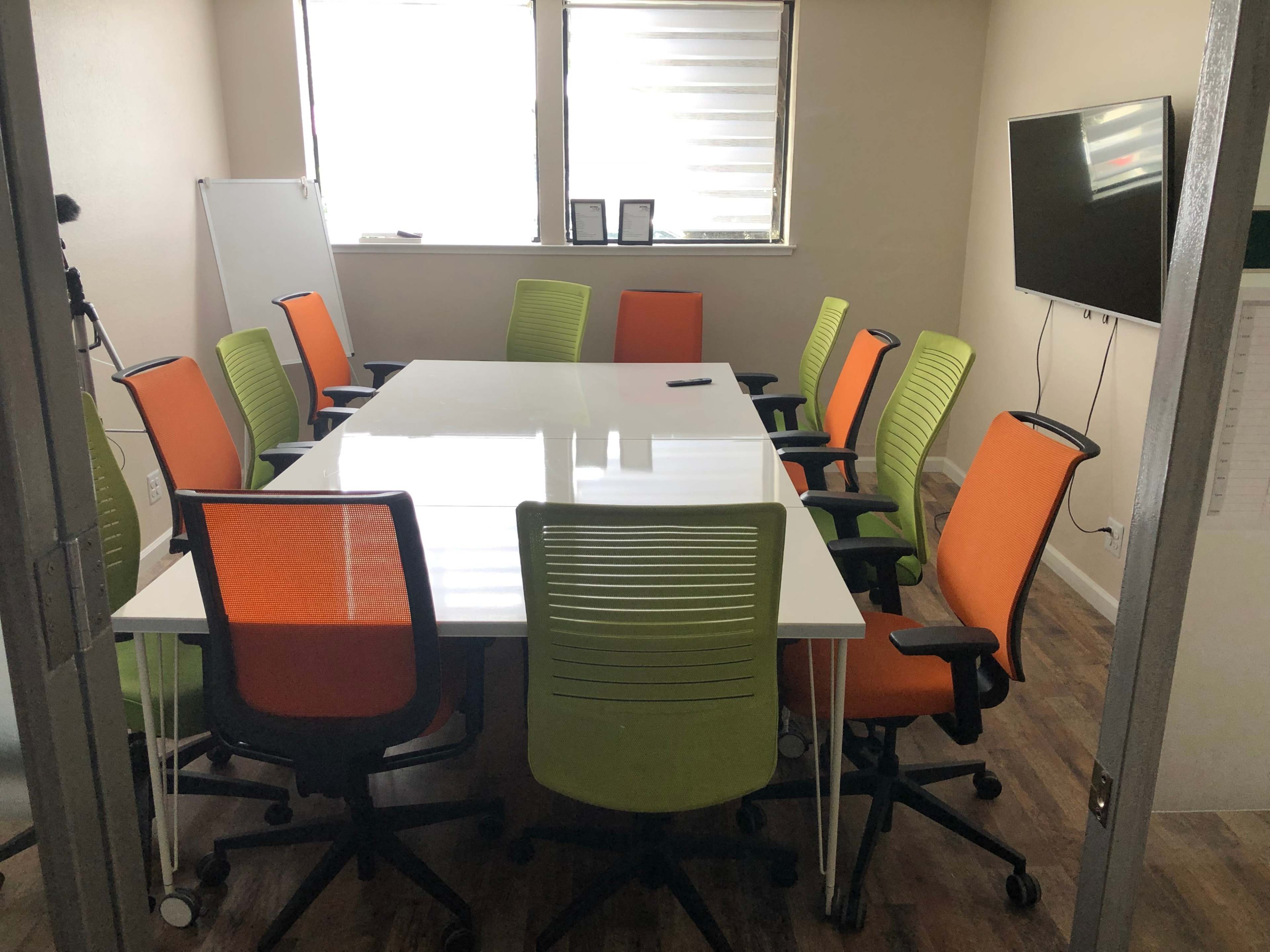 A conference room features a large white table surrounded by colorful chairs in orange and green.