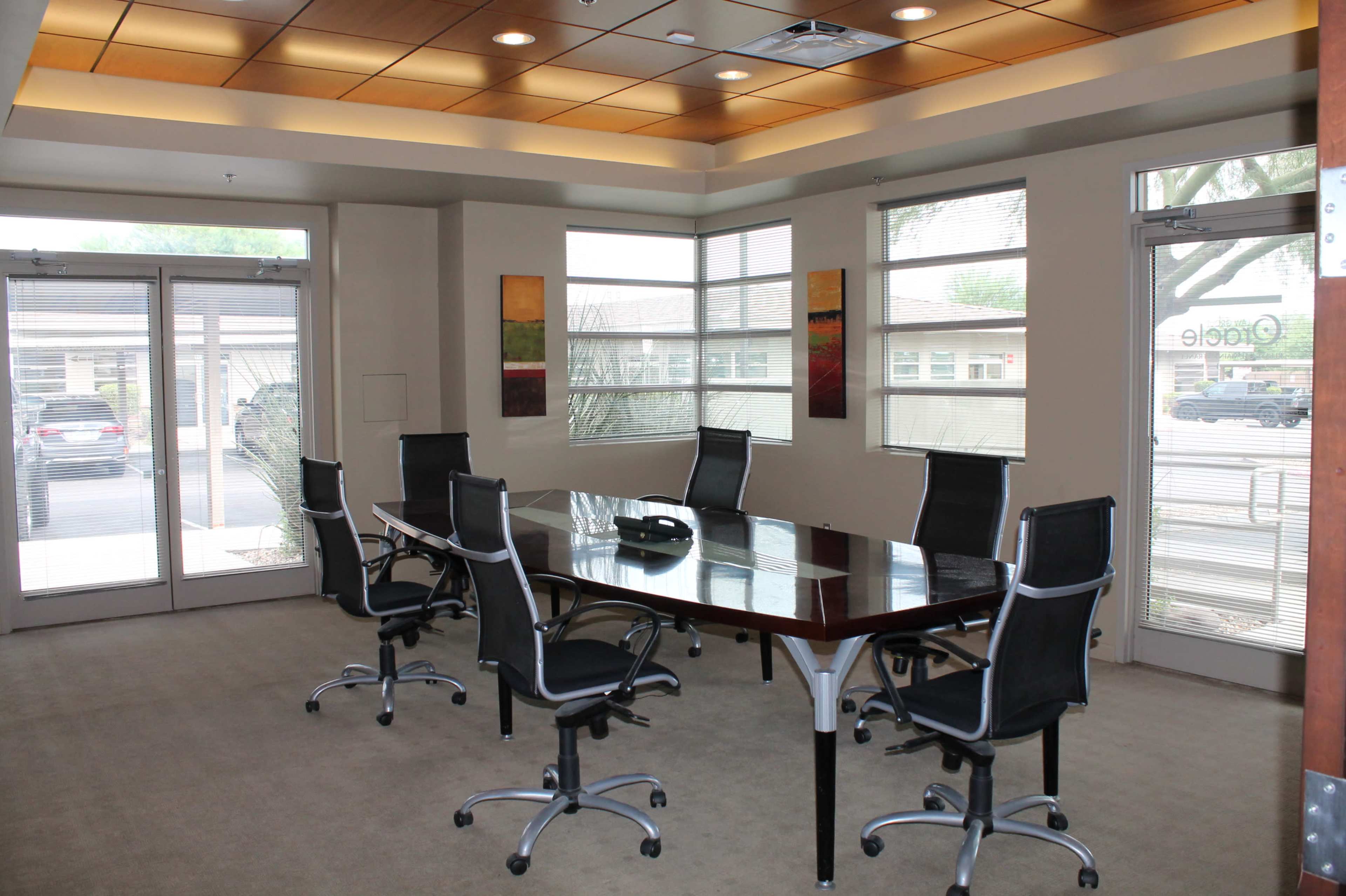 A conference room with a large wooden table surrounded by six black office chairs, featuring windows that provide natural light and a view of the exterior.