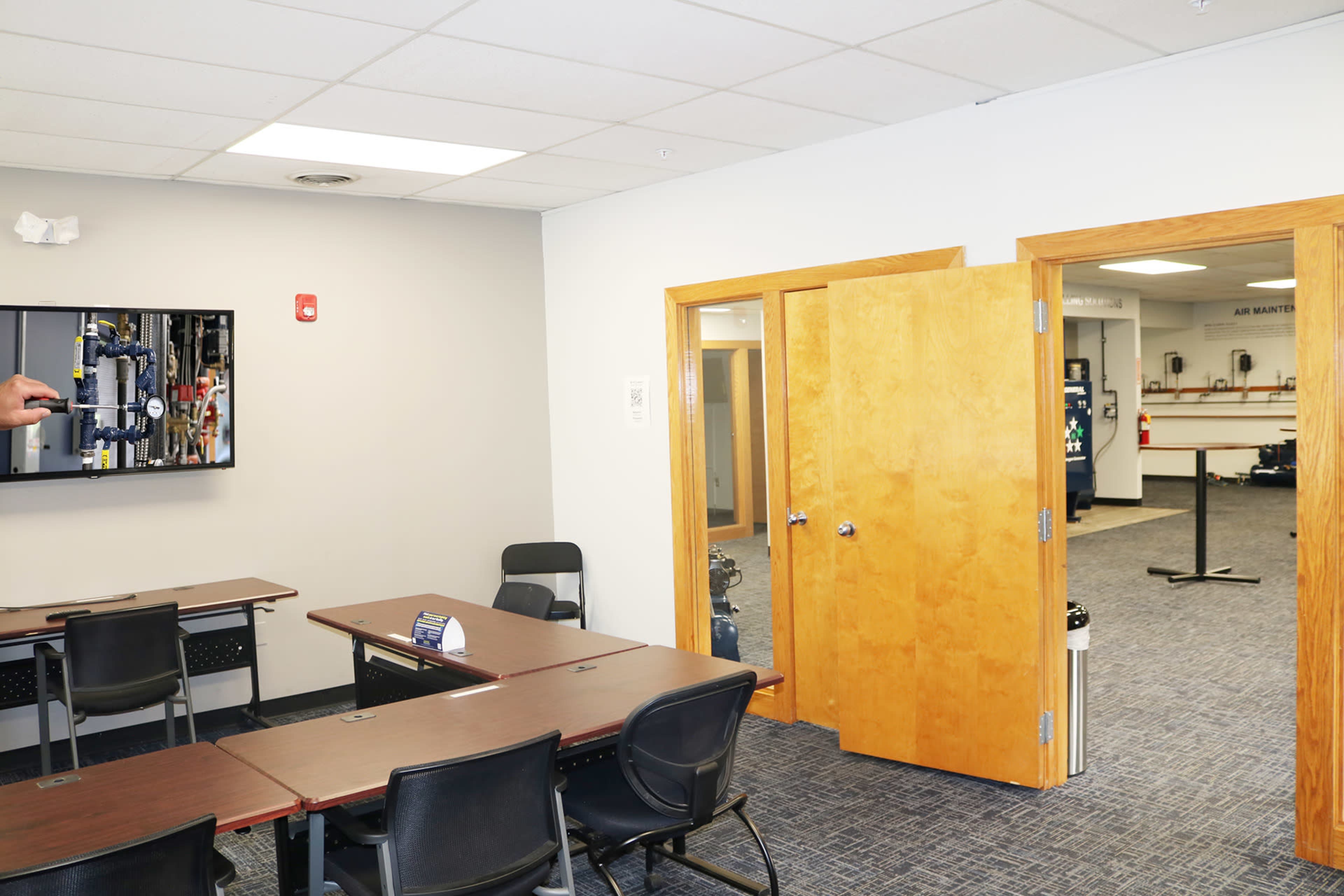 A well-lit office space featuring a large table and chairs, a wall-mounted television, and an open doorway leading to another room with equipment and tools.