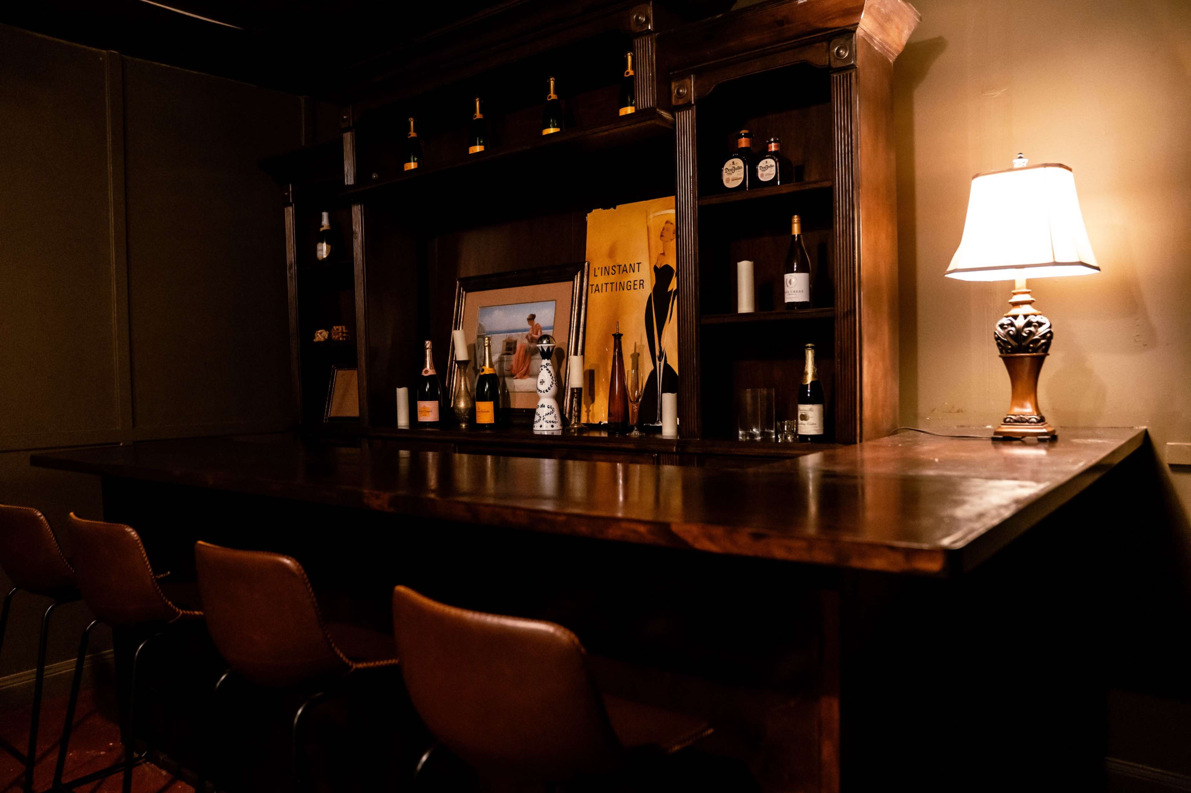 The image shows a dimly lit bar area with a wooden counter, high stools, and shelves displaying various bottles and a framed picture.