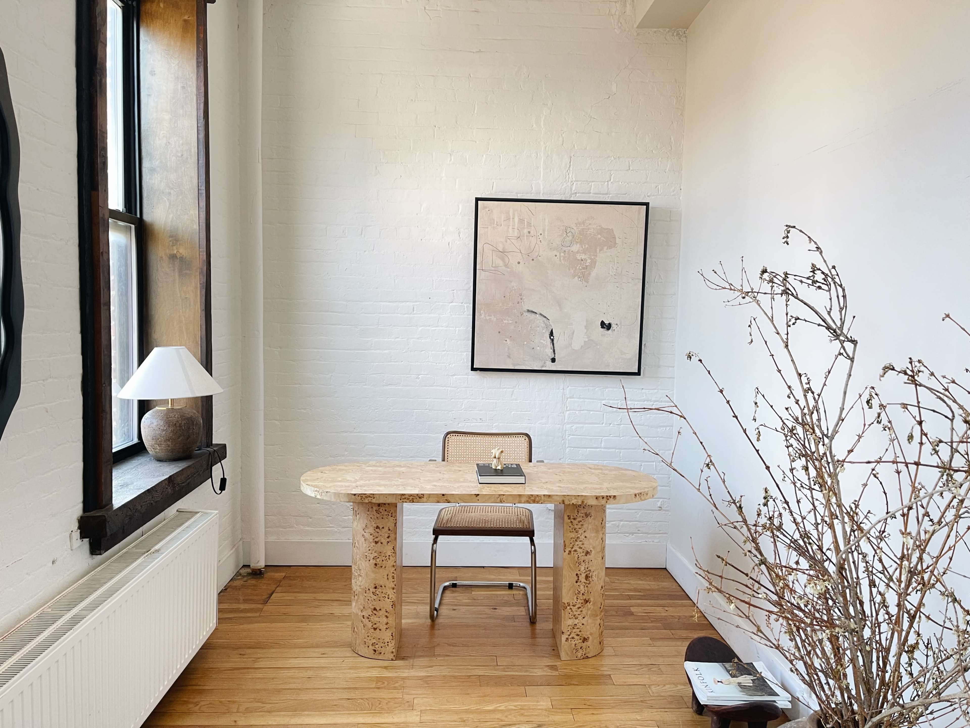 A minimalist workspace featuring a round, stone table with a chair, a small lamp, an artwork on the wall, and a decorative branch in a corner.