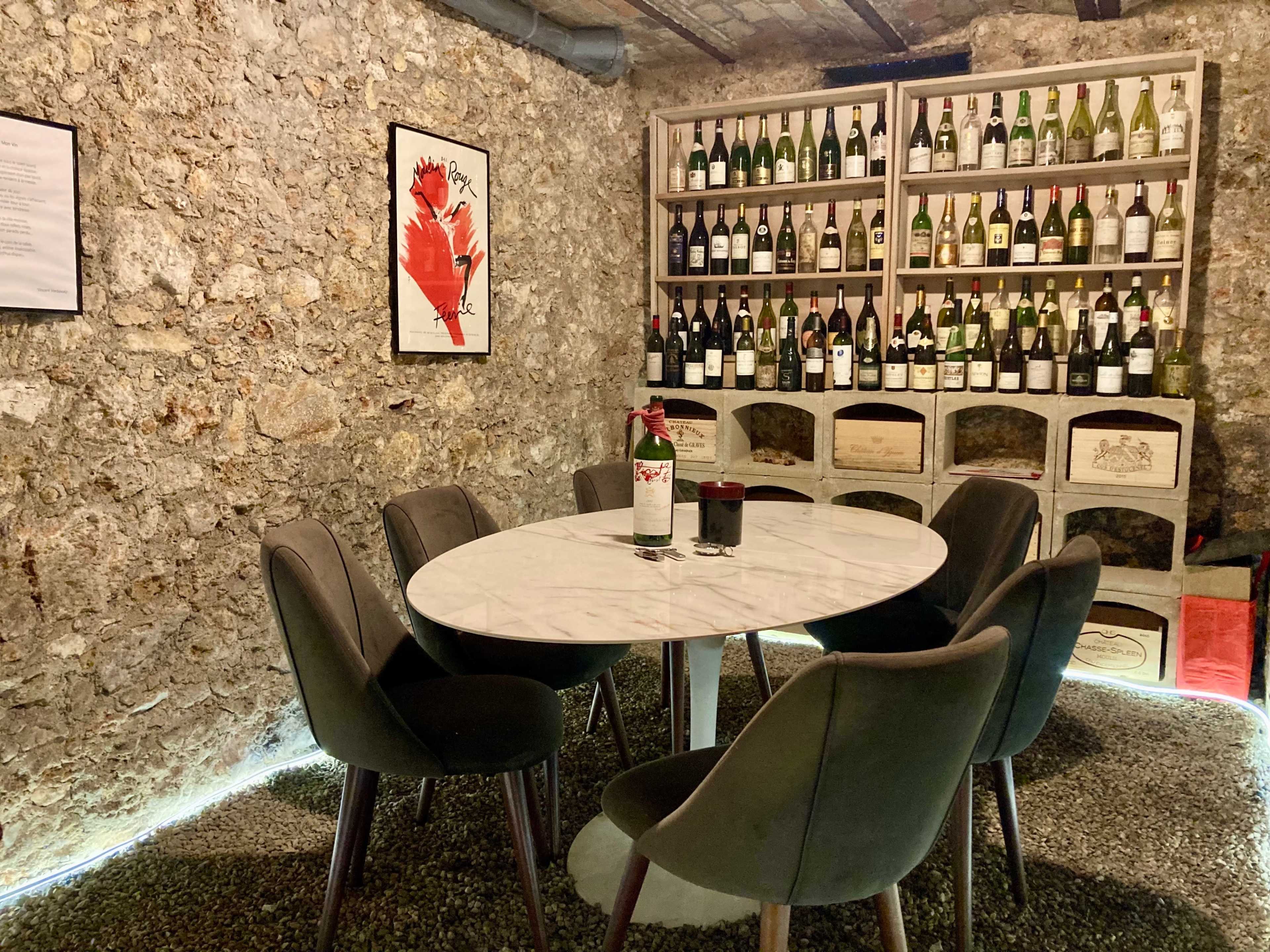 A round marble table is surrounded by six black chairs in a stone-walled room, with shelves displaying numerous bottles of wine in the background.