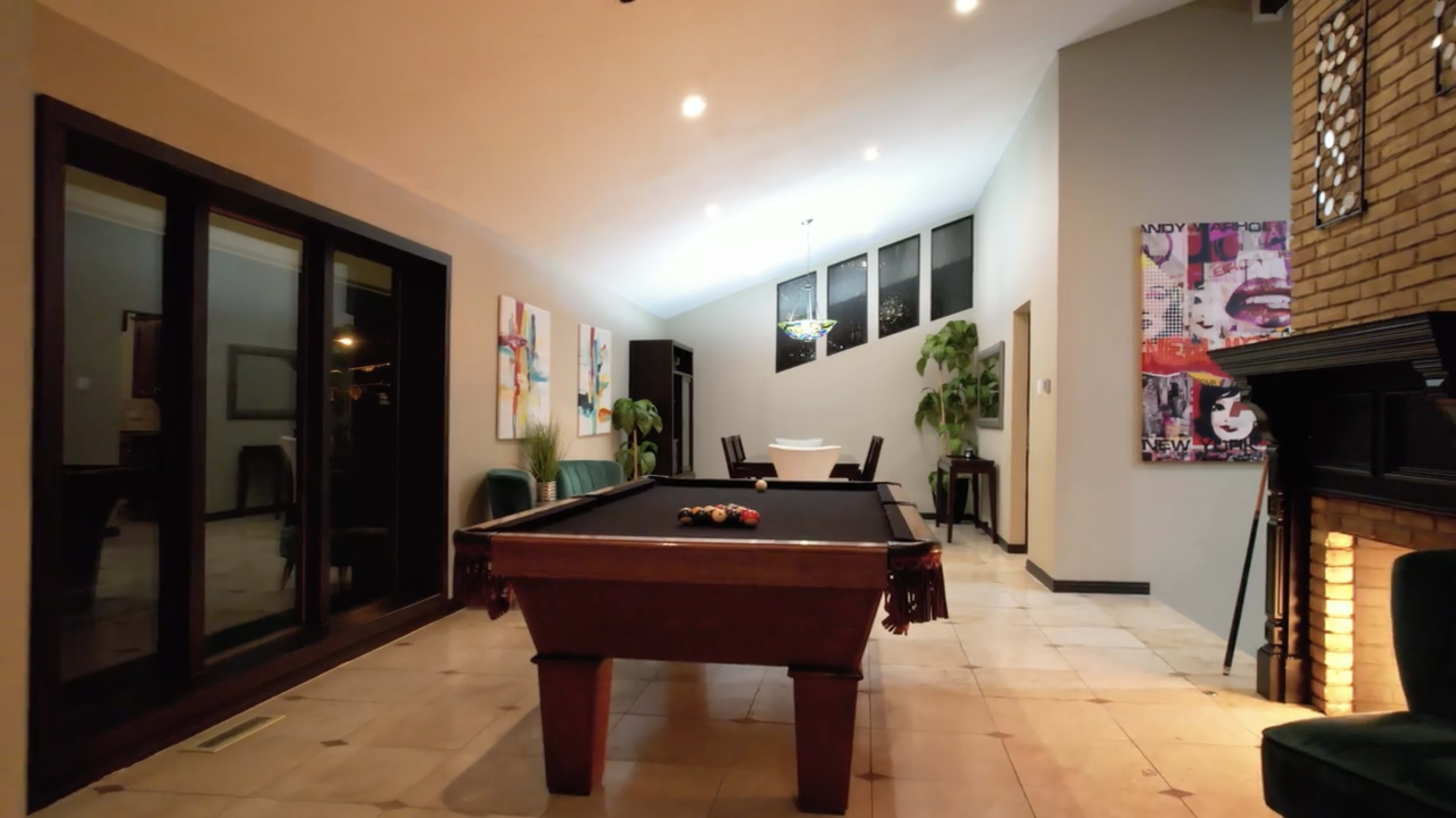 A spacious room with a pool table in the center, large windows letting in light, and various pieces of artwork on the walls.