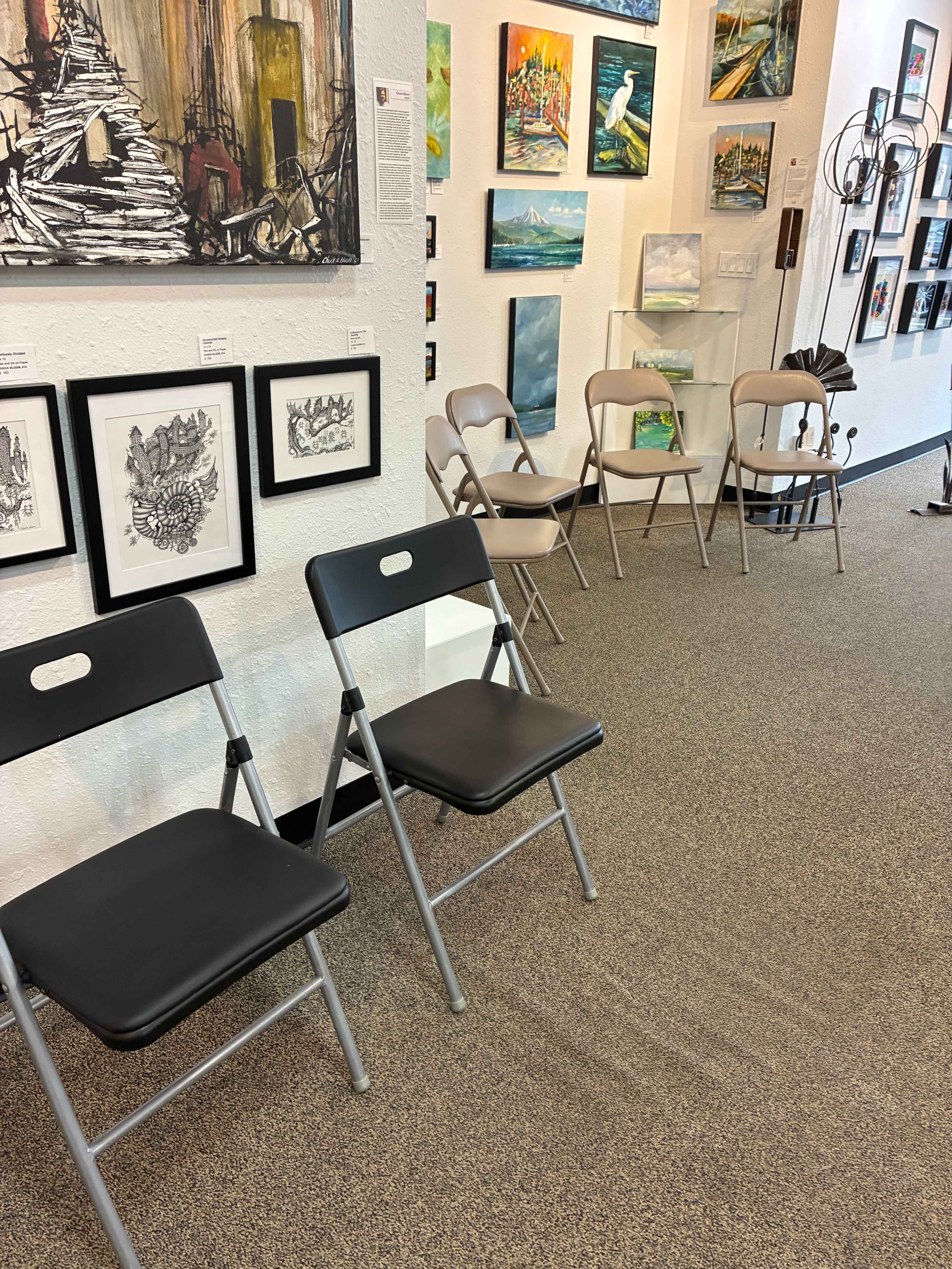 The image shows a gallery space with several pieces of artwork on the walls and a row of folding chairs along one side.