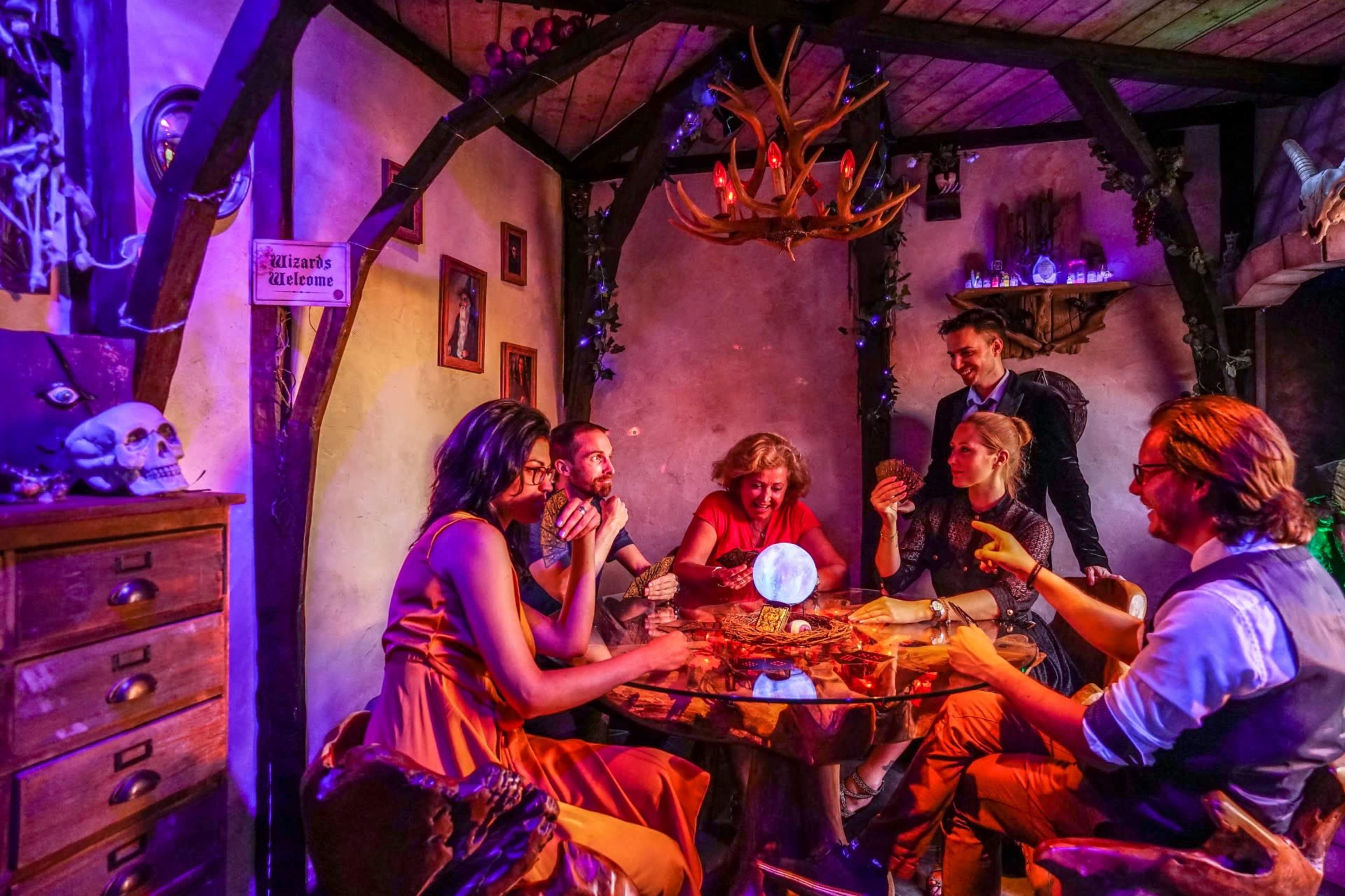 A group of five people sits around a table in a dimly lit room with mystical decor, engaged in a séance, with a crystal ball at the center.