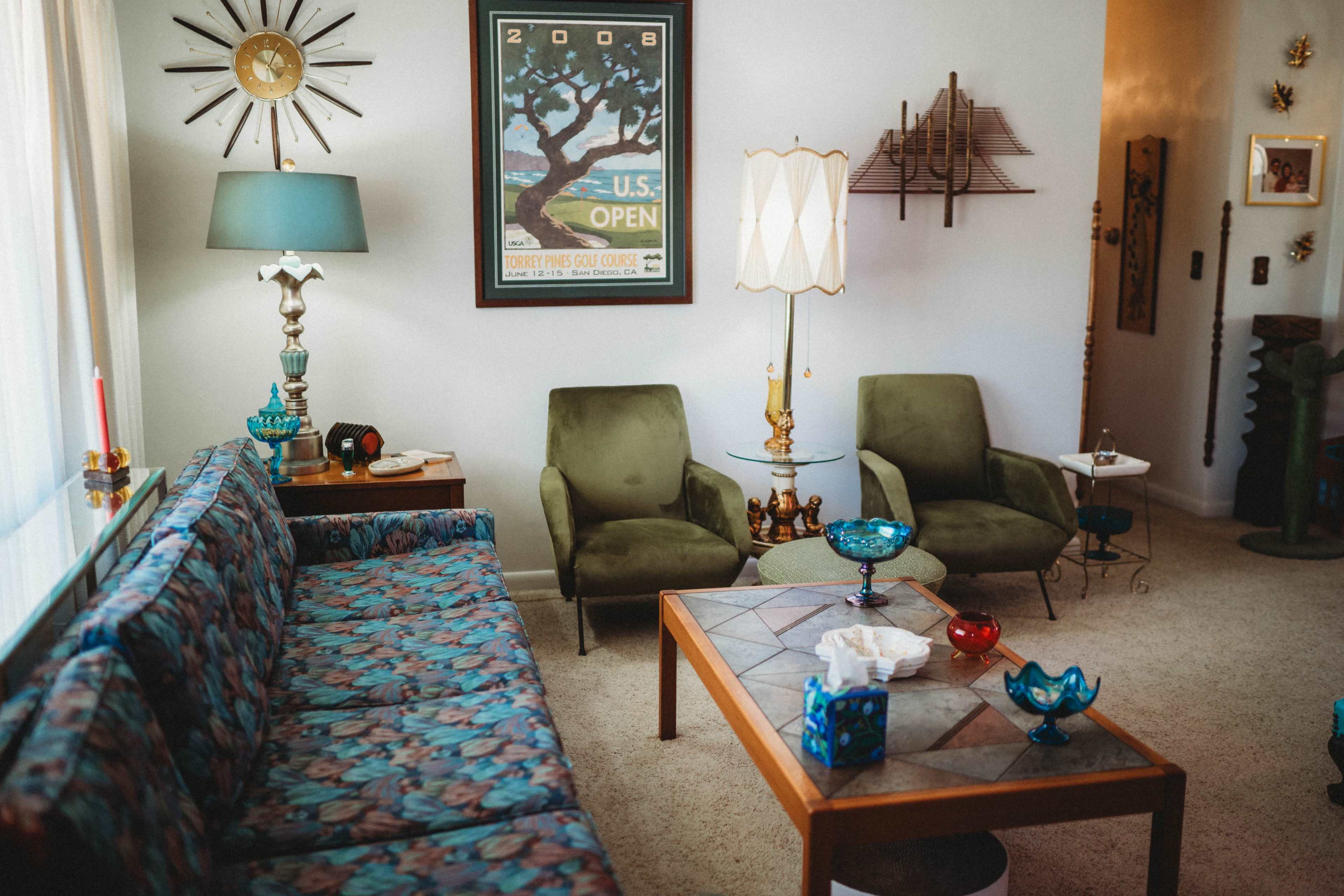 The image shows a vintage living room with a patterned sofa, two green armchairs, and a wooden coffee table adorned with decorative items.