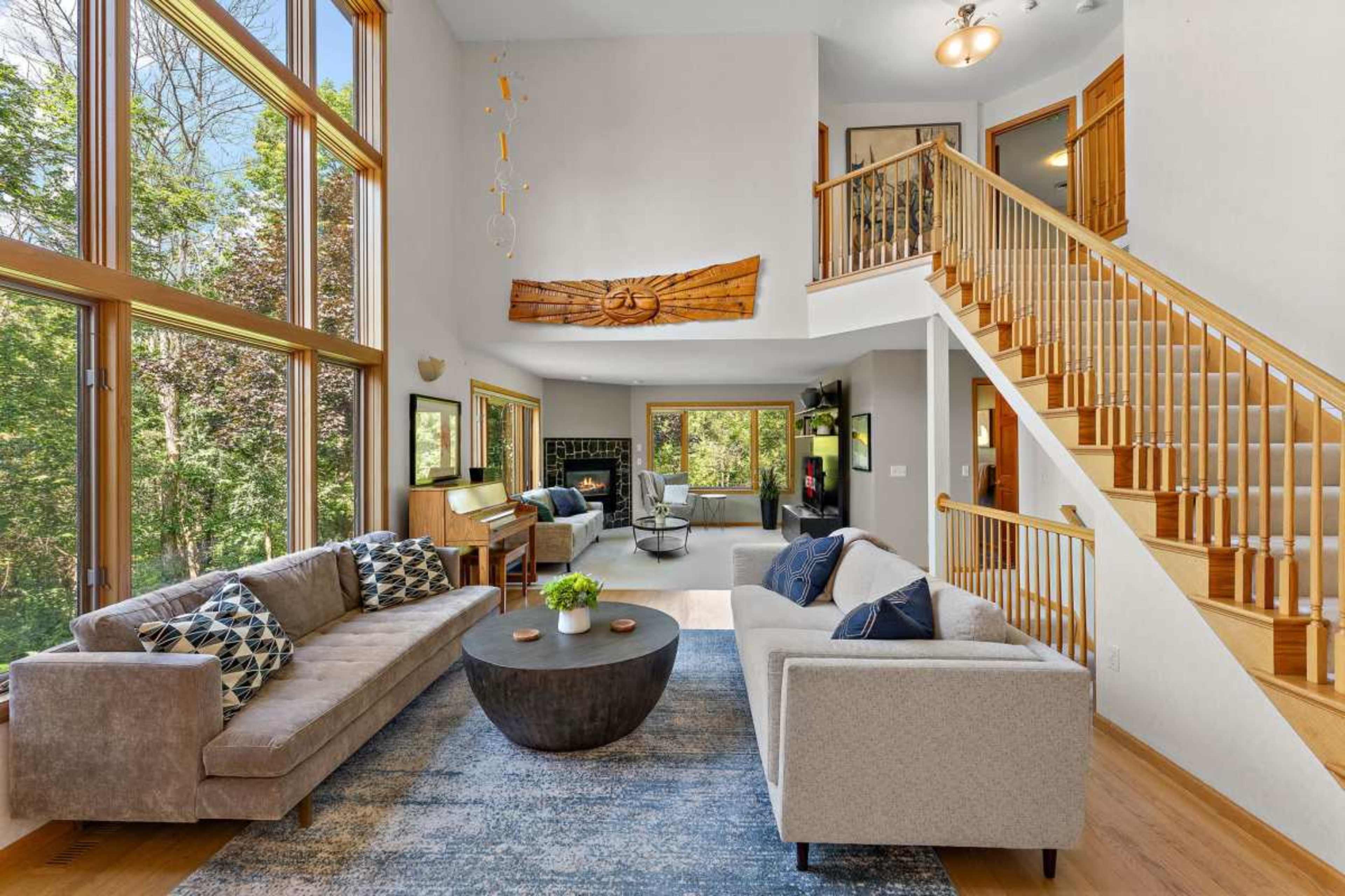 The image shows a spacious living room with large windows, a round coffee table, two sofas, and a staircase leading to an upper level.