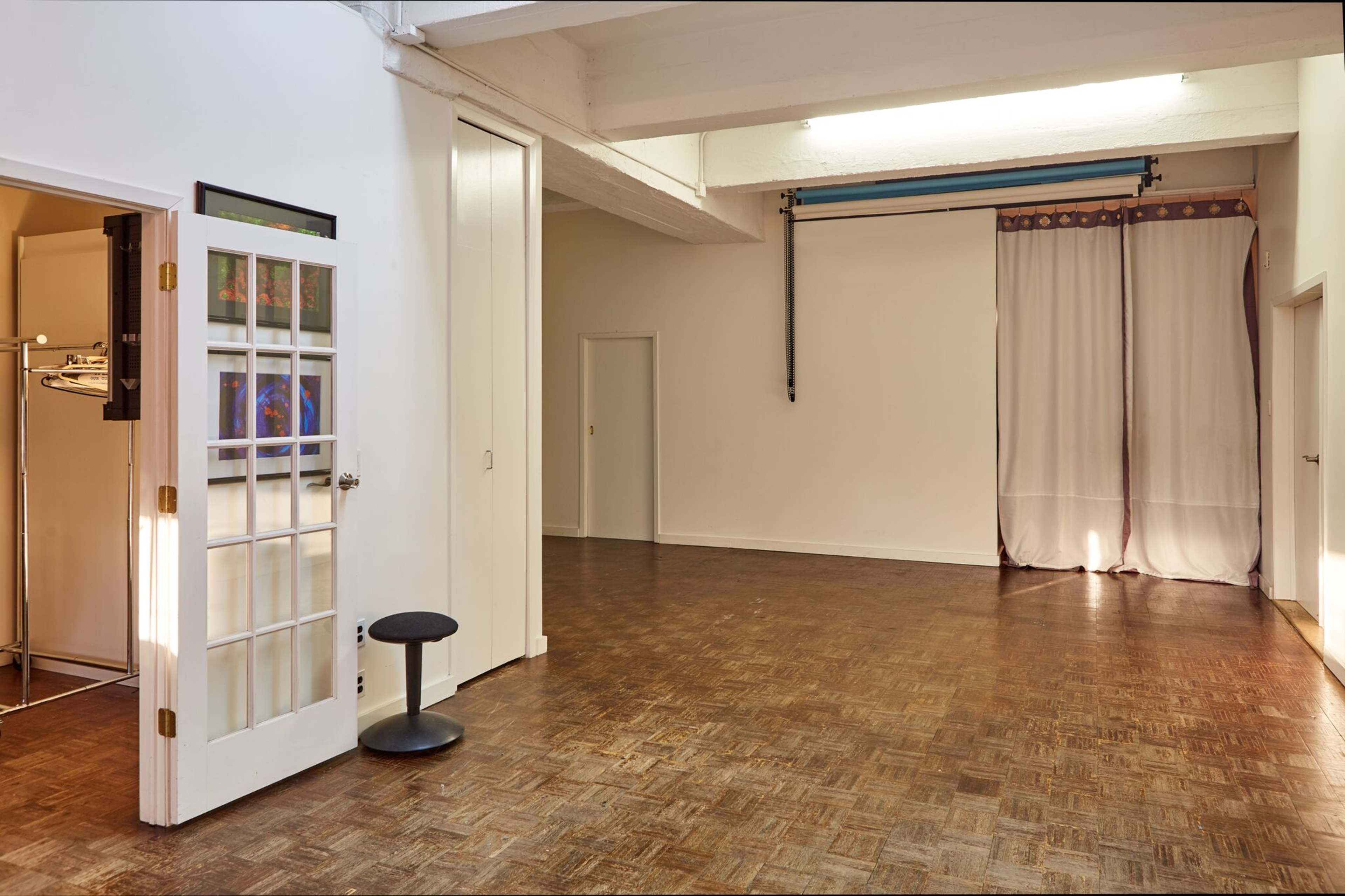 A spacious, well-lit room with polished wooden floors, featuring a door with glass panes, a small black stool, and a curtain hanging against a wall.