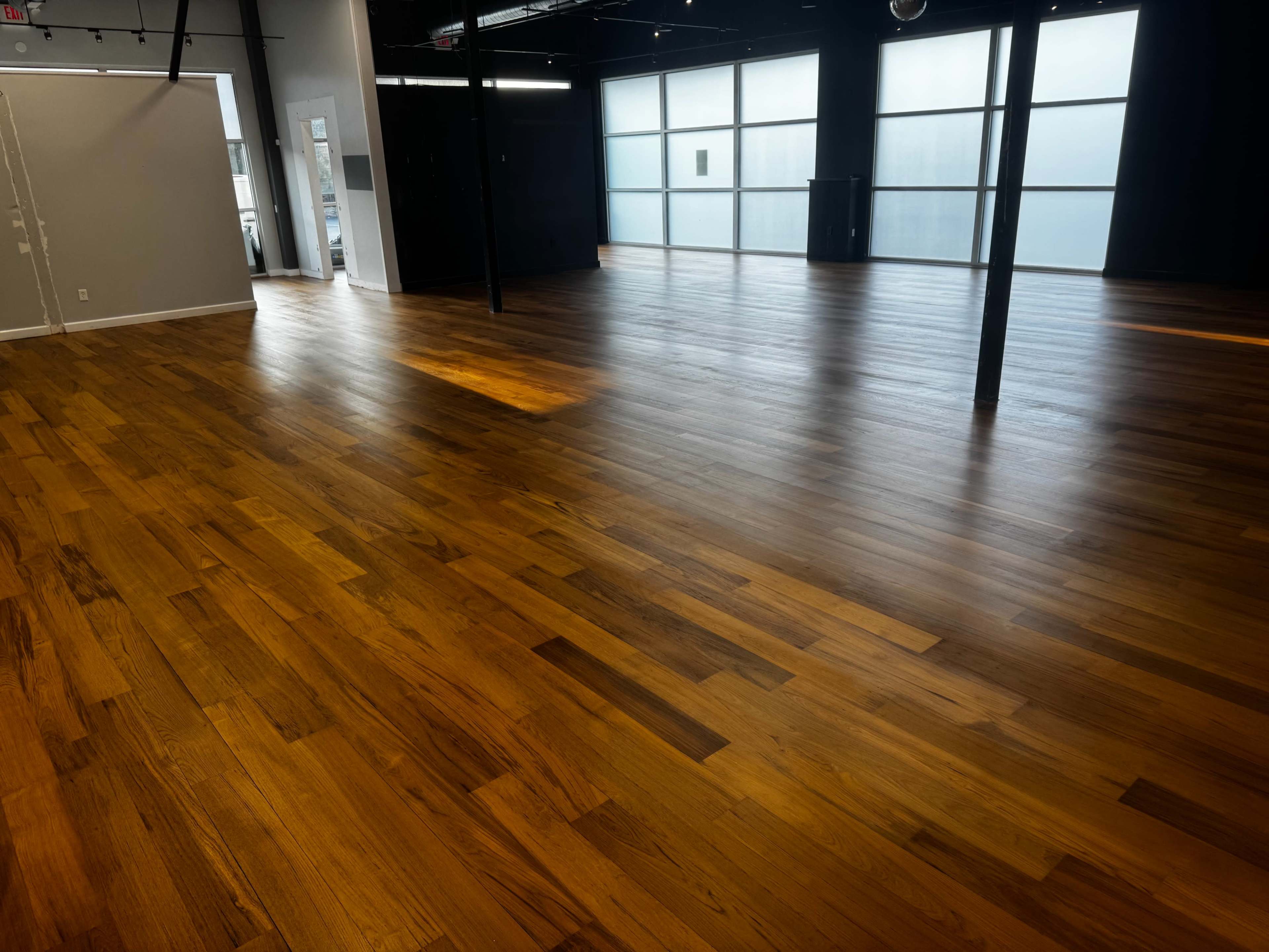 The image shows a spacious room with polished wooden floors and large windows that allow natural light to enter.