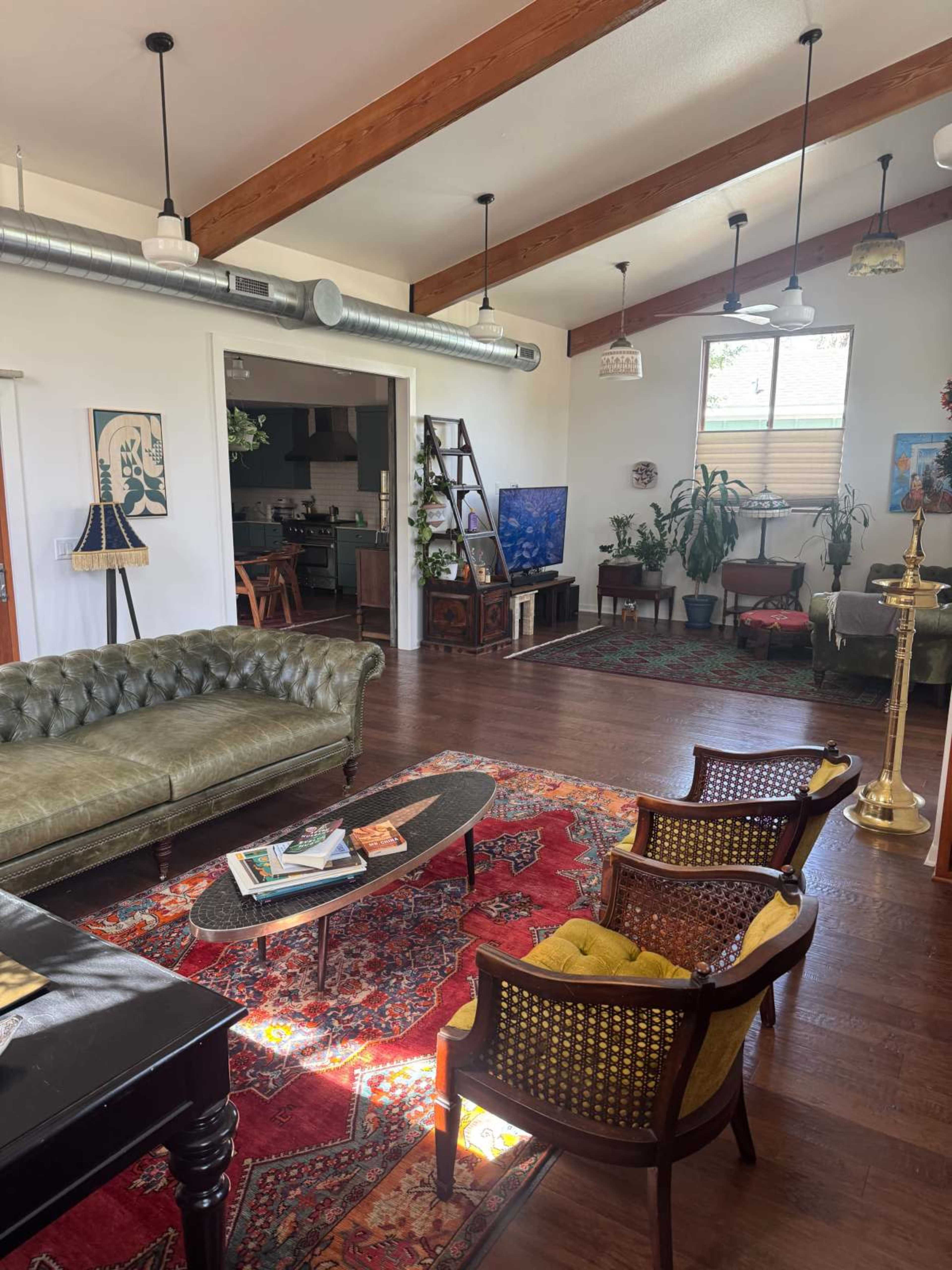 A spacious living area features a green leather sofa, two armchairs, a coffee table, a large area rug, and various plants, with a kitchen visible in the background.