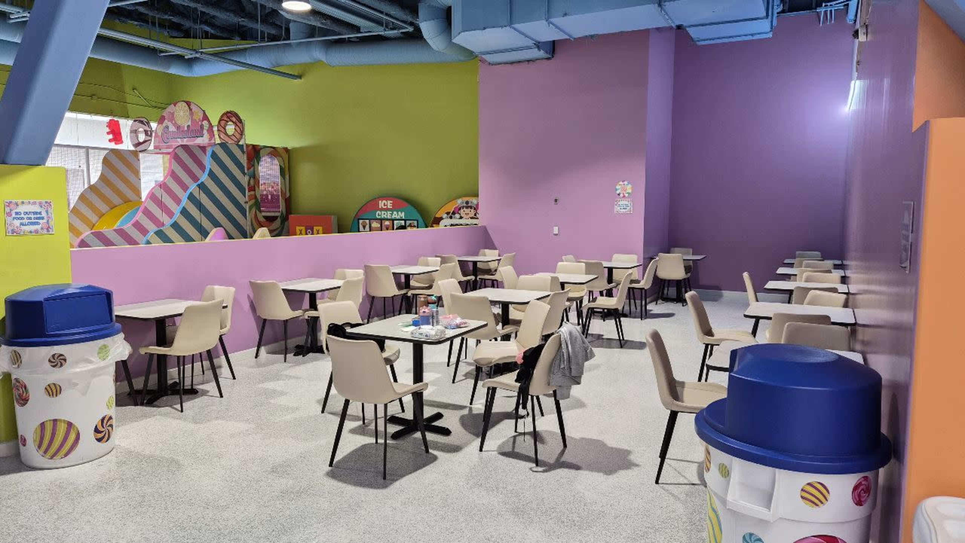 The image shows a brightly colored dessert shop with empty tables and chairs arranged in a spacious layout, featuring walls painted in pastel colors.