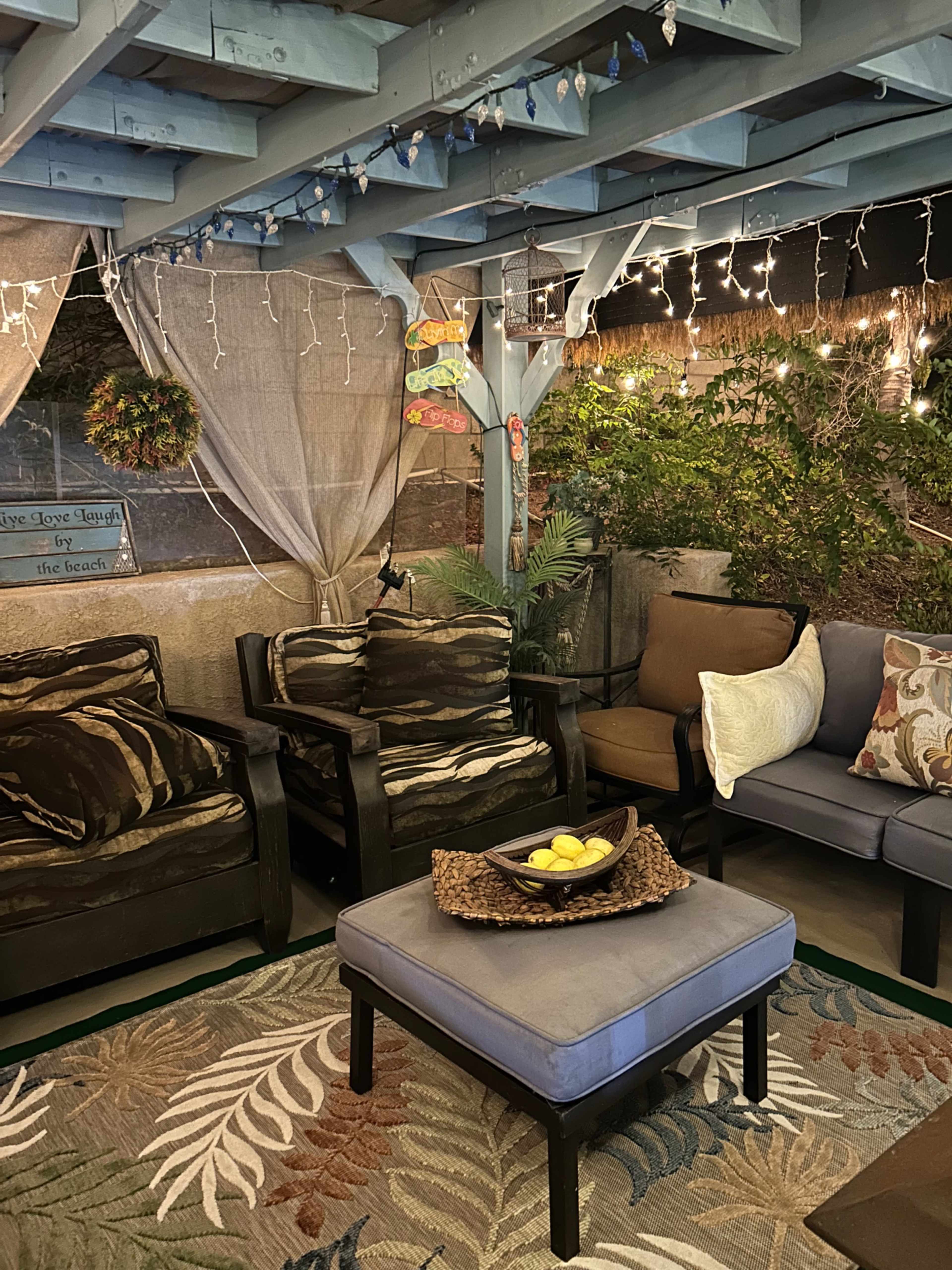 The image shows a cozy outdoor seating area illuminated by string lights, featuring a mix of cushioned chairs and a table with a bowl of lemons.