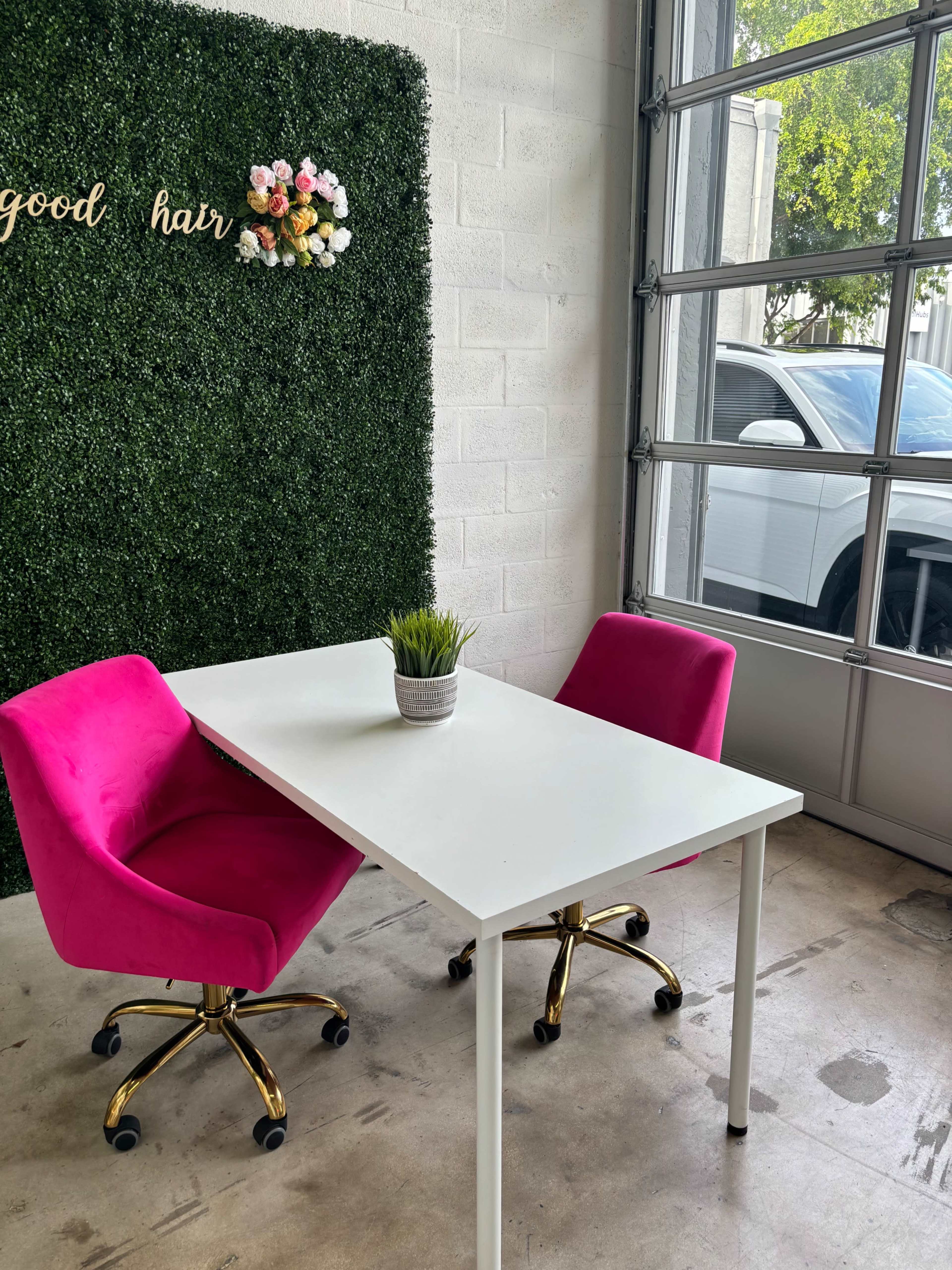 A small white table with two pink chairs sits in front of a greenery wall adorned with flowers and the words "good hair."