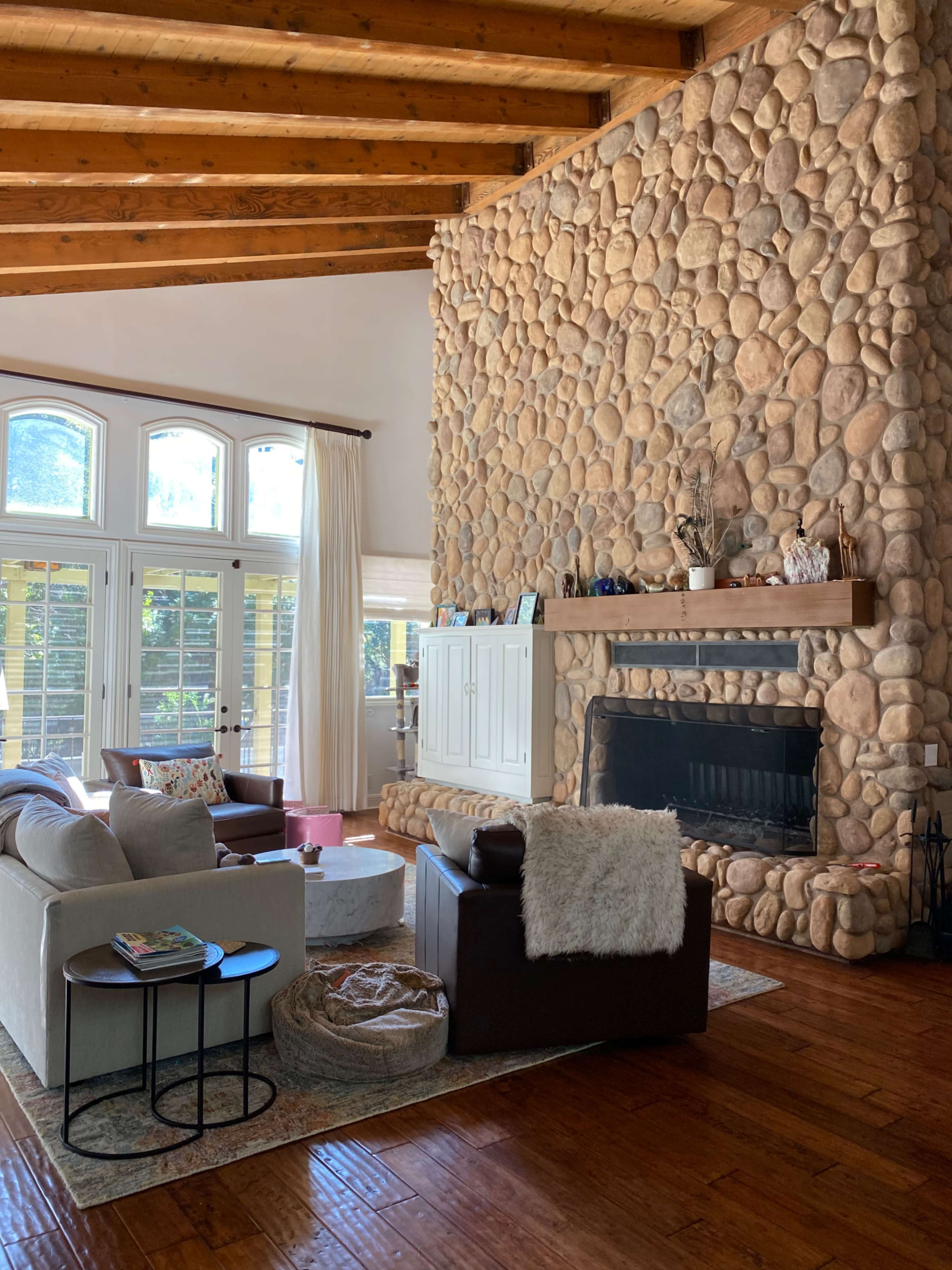 The image shows a spacious living room featuring a large stone fireplace, wooden beams on the ceiling, and comfortable seating arrangements.