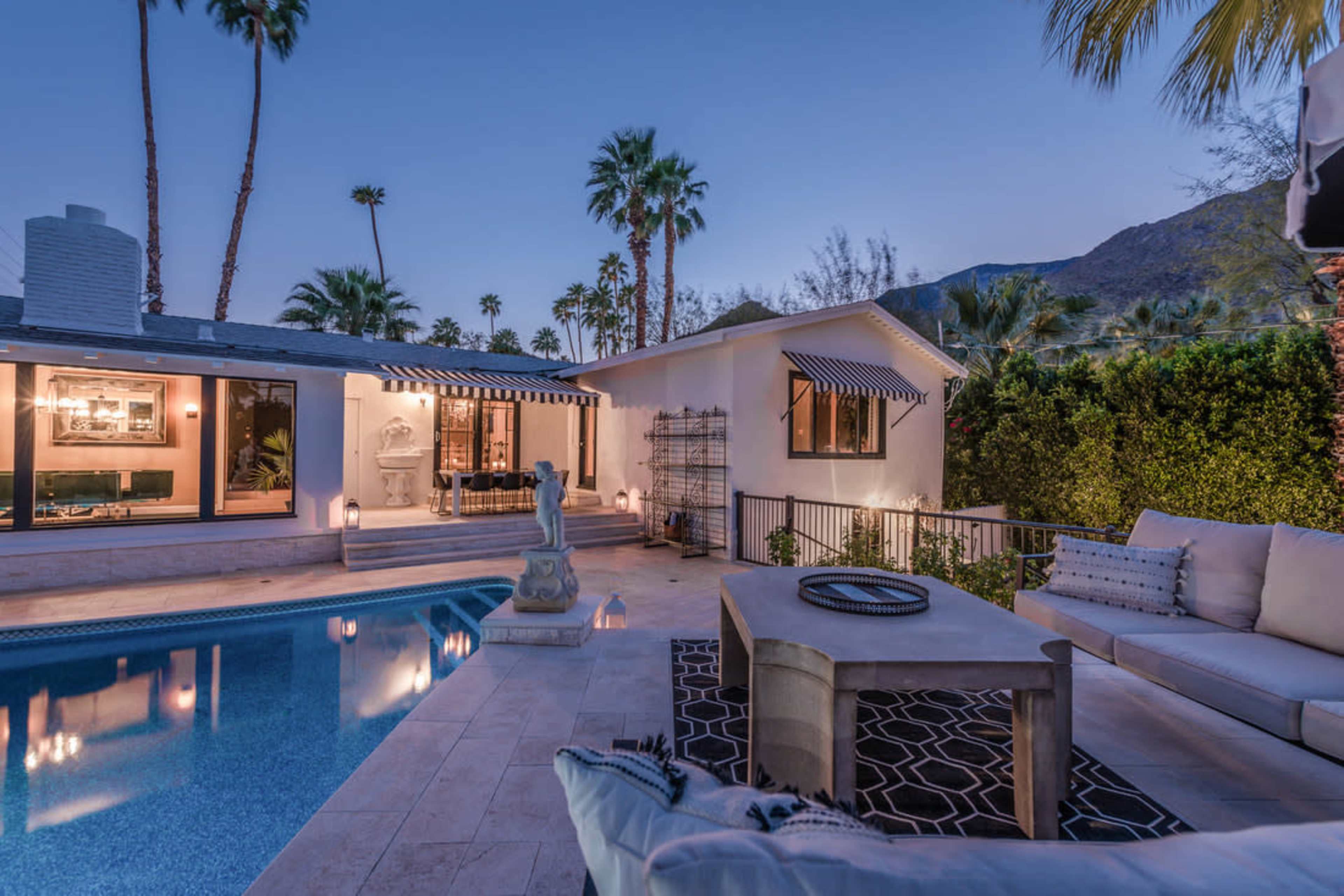 A serene outdoor space featuring a swimming pool, a table, and lounge seating, surrounded by palm trees and mountains under a twilight sky.
