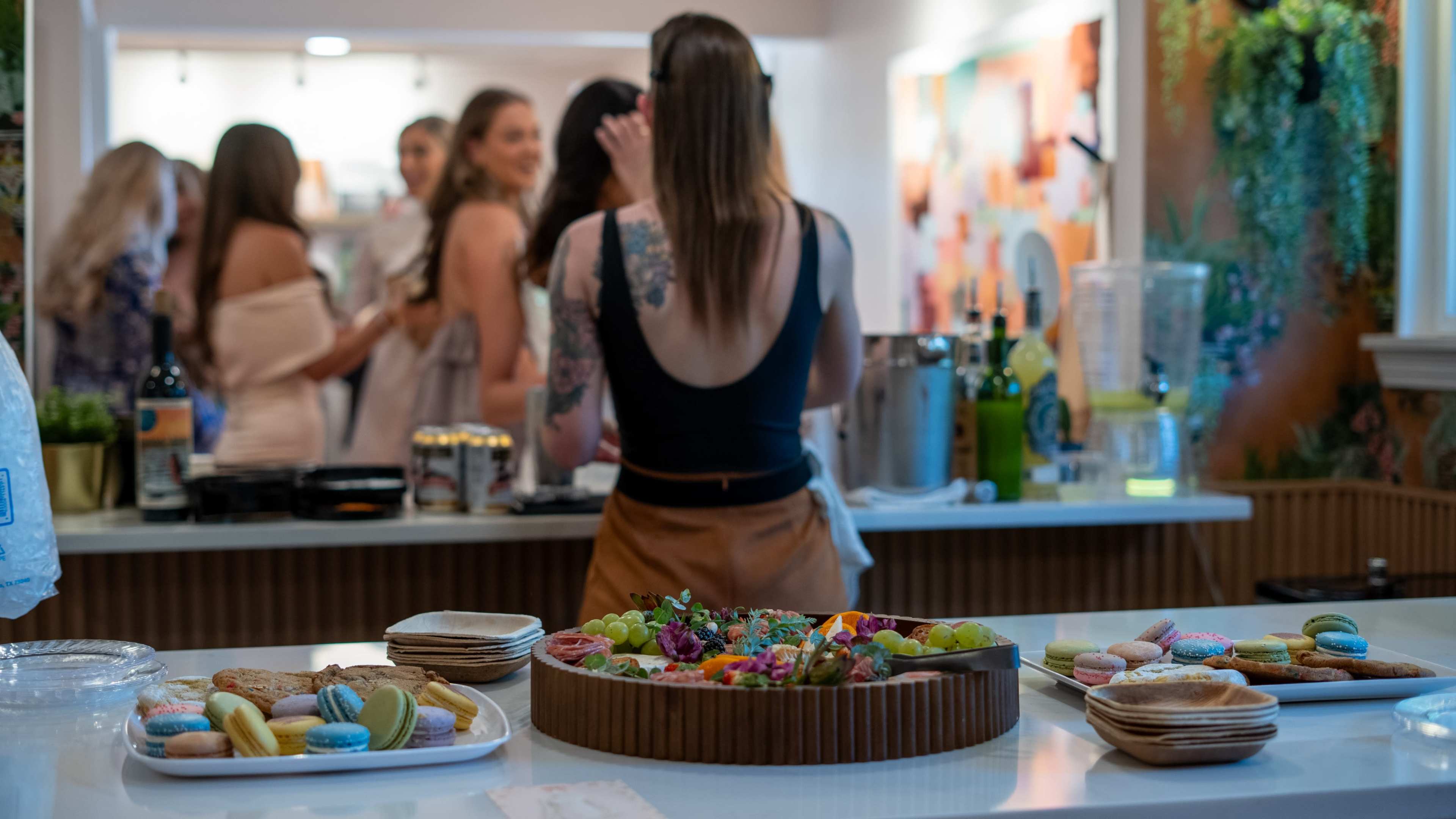 A person stands in front of a table displaying a variety of snacks, including macaroons and a fruit platter, while a group of people chats in the background.