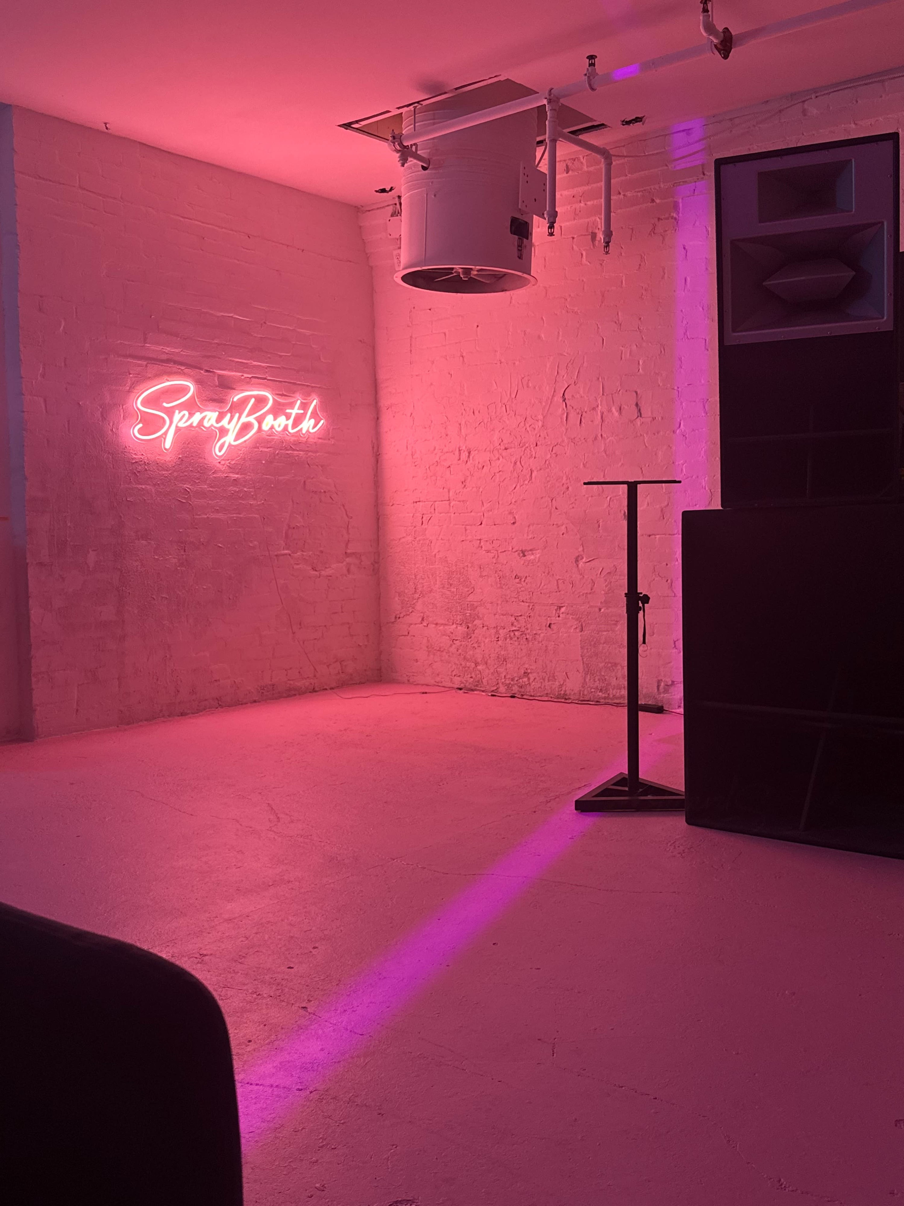 The image shows a minimalist interior space with pink lighting, featuring a neon sign that reads "SprayBooth," a tall speaker, and a small table.