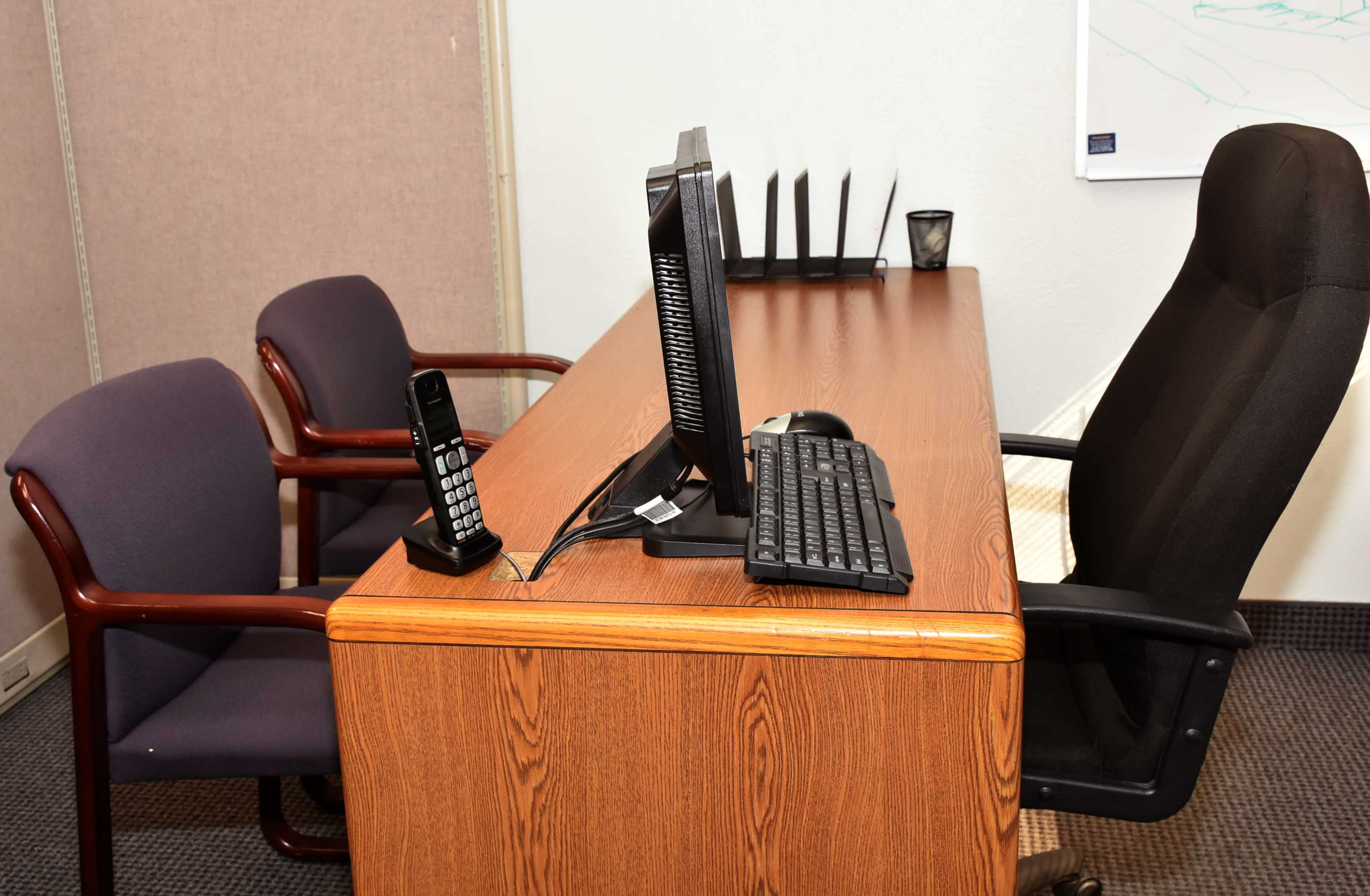A wooden desk with a computer monitor, keyboard, a telephone, and two chairs is set up in an office space.