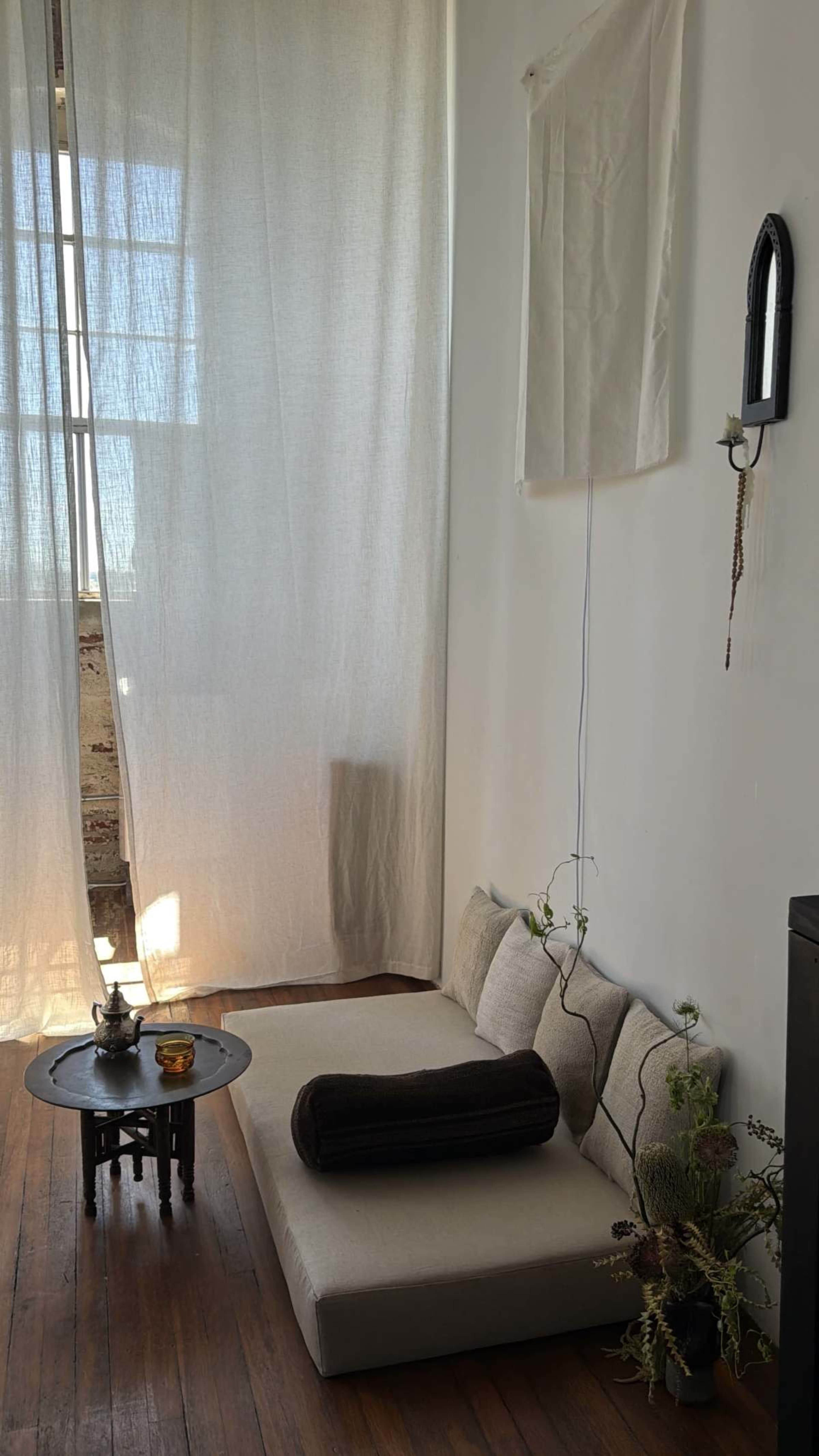 The image shows a cozy corner of a room with a low seating area, a small round table, and sheer curtains letting in soft light.