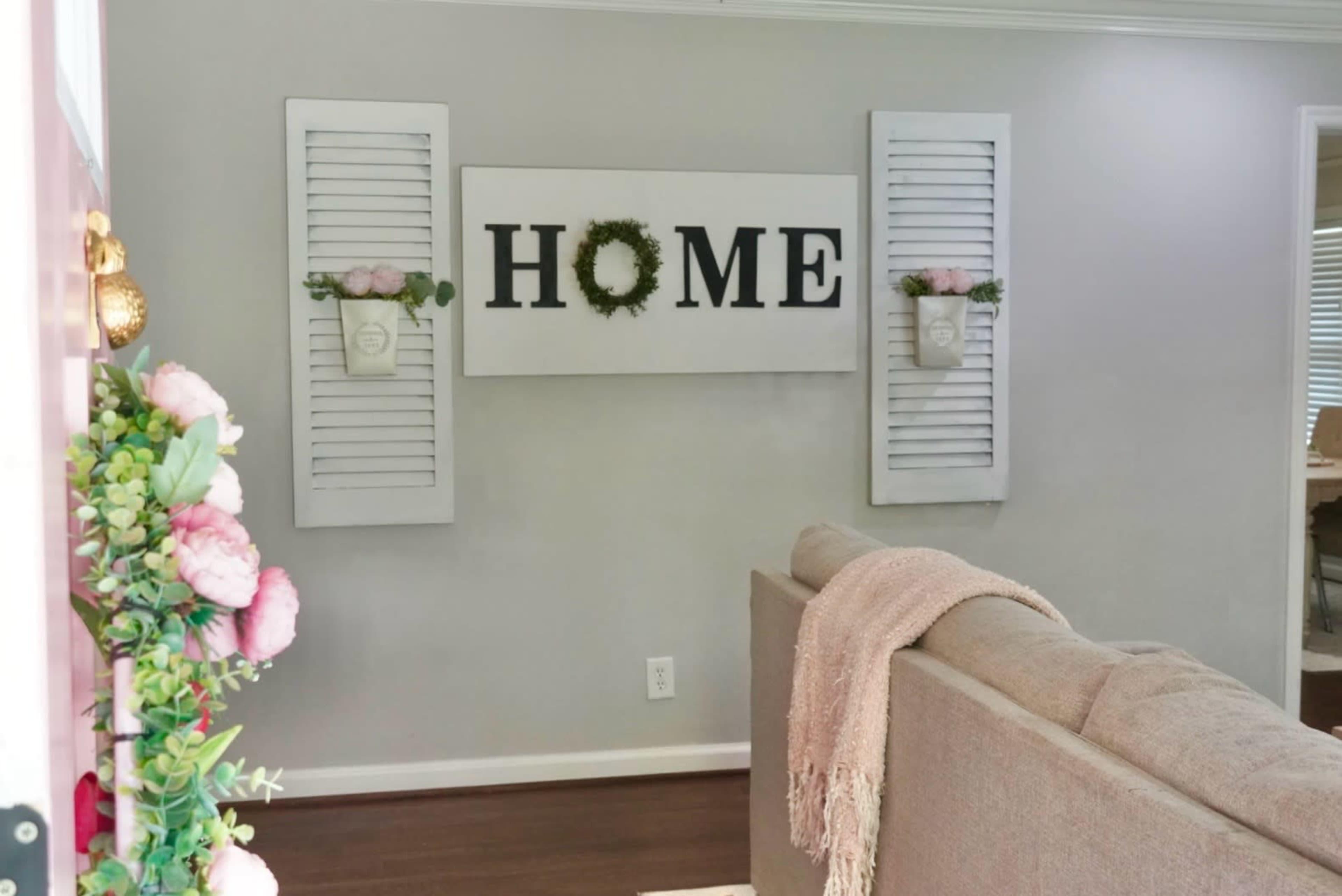 A living room features a beige sofa with a decorative throw, surrounded by wall decor that includes a sign reading "HOME" and floral arrangements in wall-mounted planters.