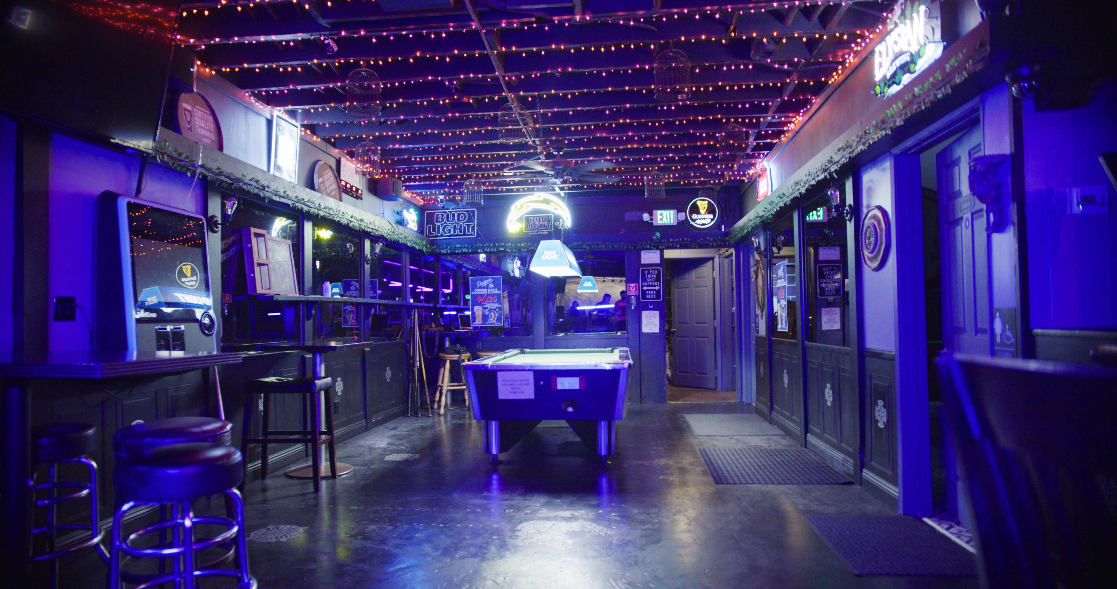 An empty bar interior with a pool table, illuminated by purple lights and decorative string lights along the ceiling.