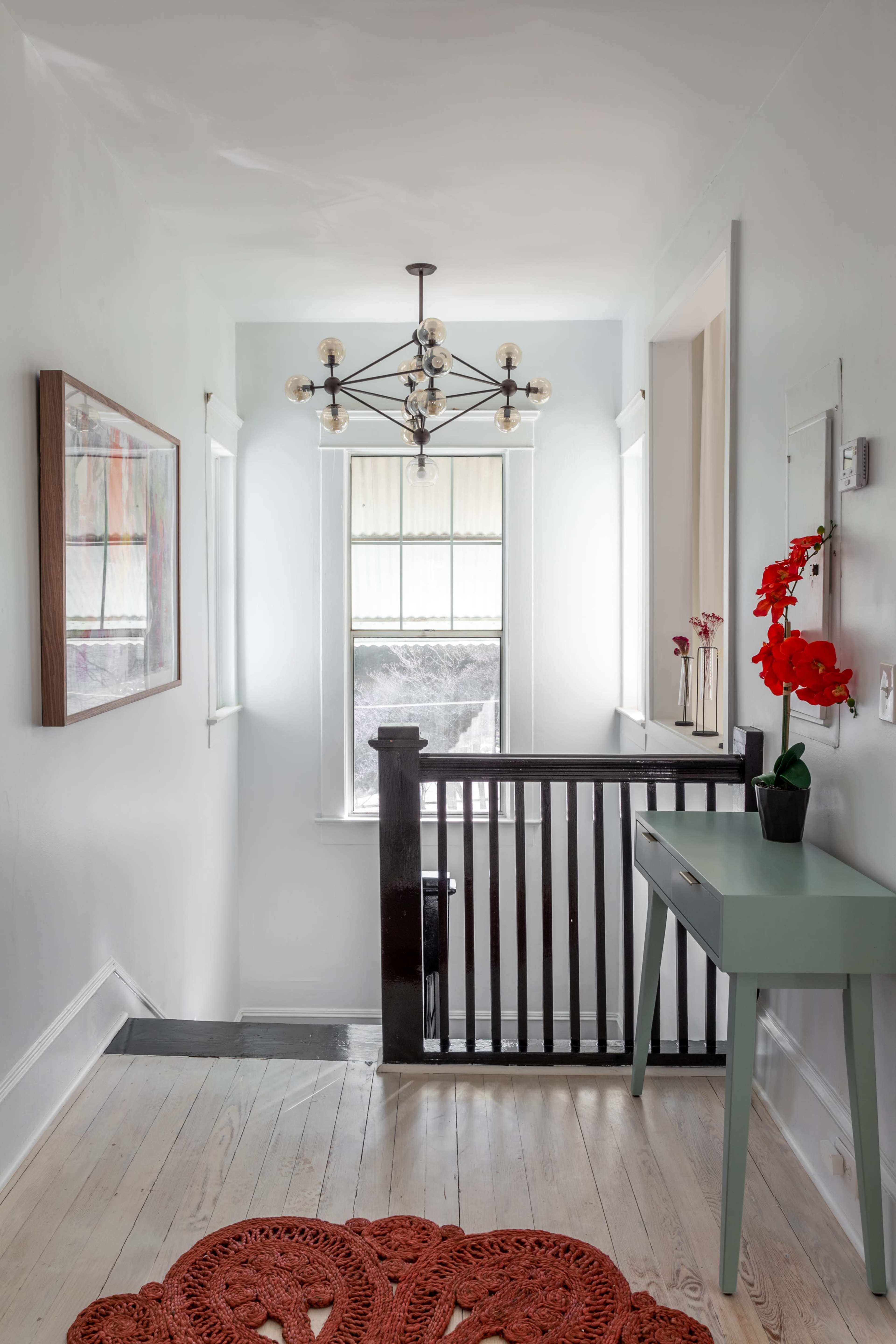 A narrow hallway with a modern chandelier, two windows, a small table, and a vibrant red rug.