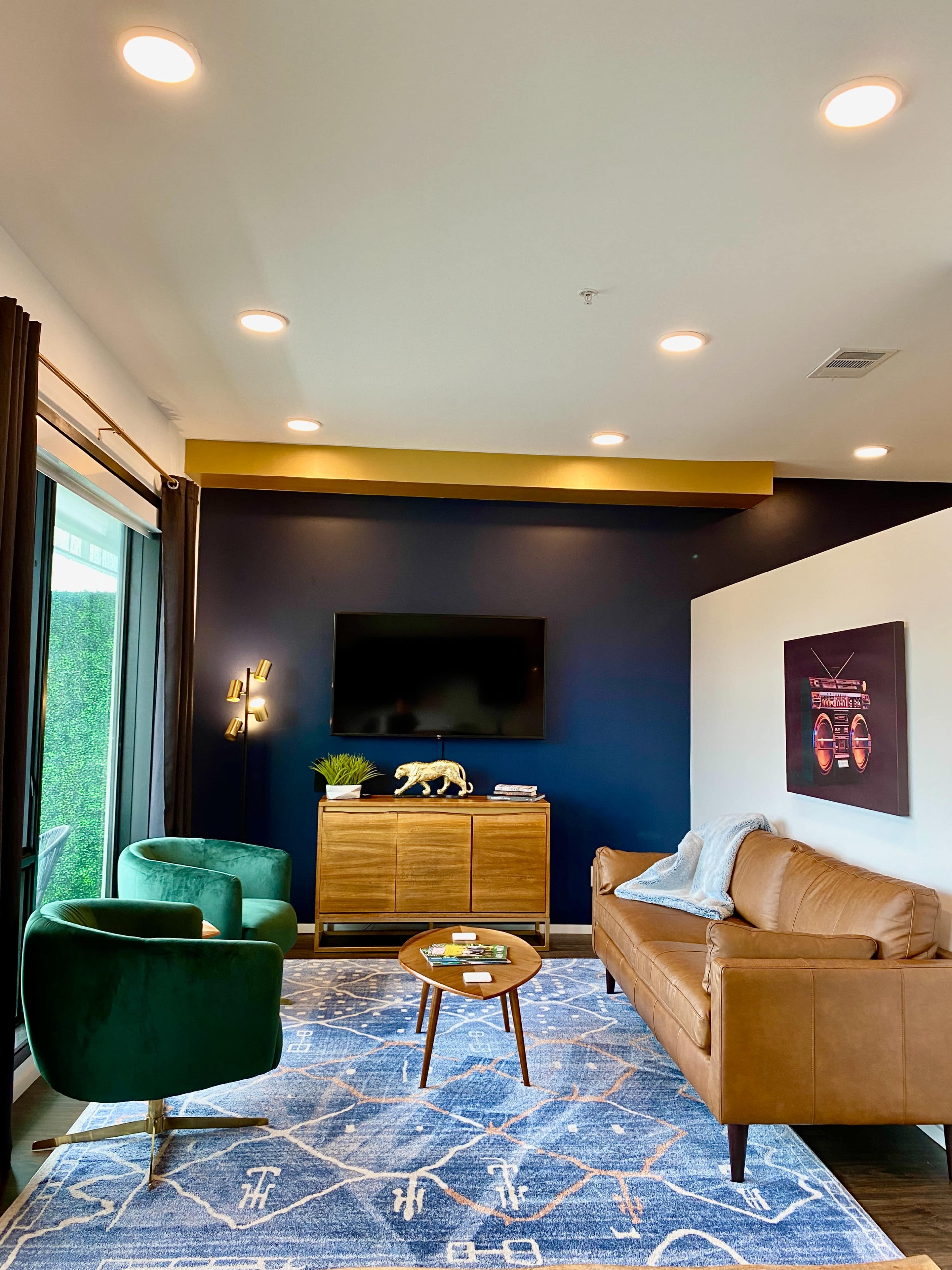 A modern living room features a brown leather sofa, two green armchairs, a wooden cabinet, a round coffee table, and a mounted television against a dark blue wall.