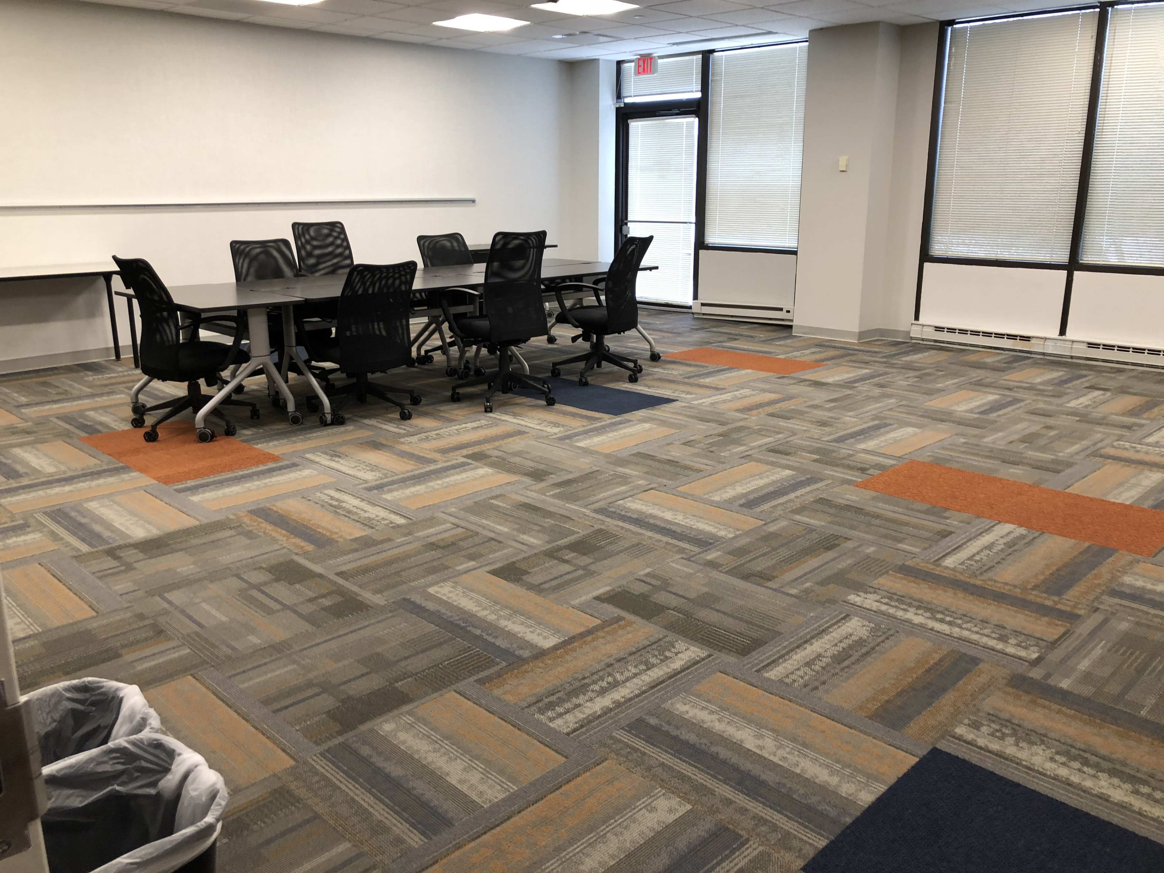 An empty conference room features a large table surrounded by black chairs on a patterned carpet with sections of orange and gray.