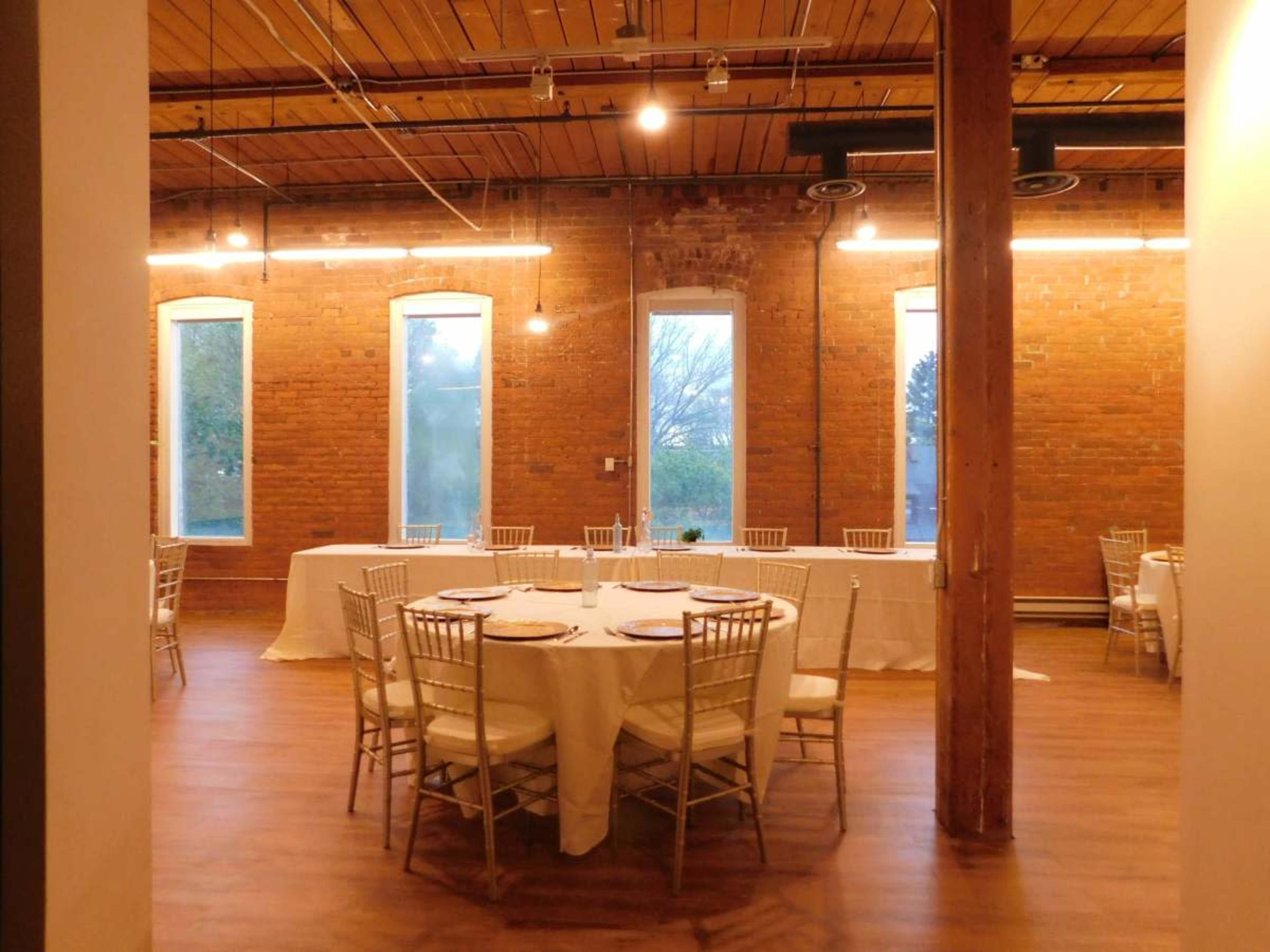 A banquet hall with round tables covered in white tablecloths and gold chairs, set against a backdrop of brick walls and large windows.