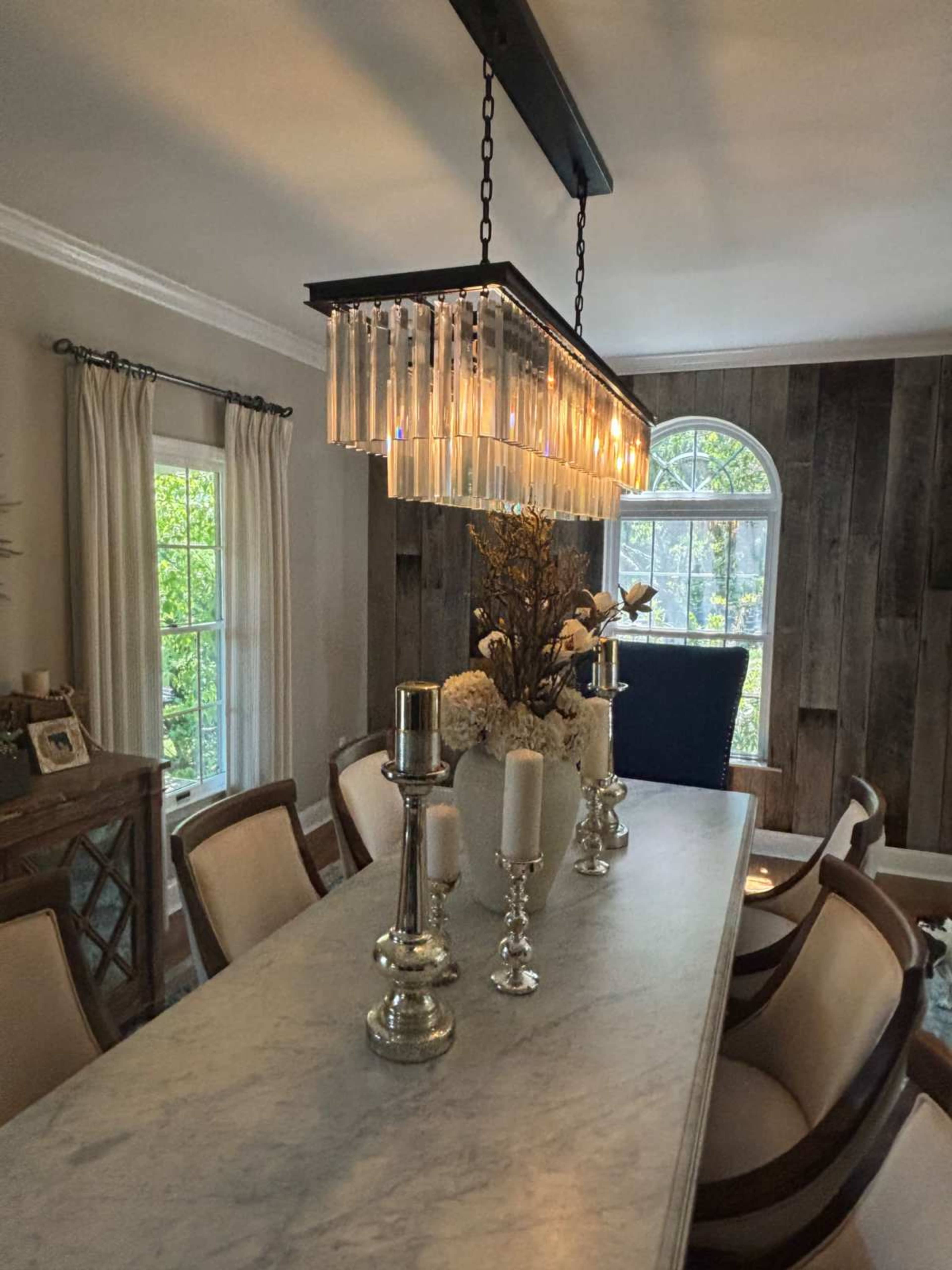 A modern dining room features a long marble table with elegant chairs, adorned with tall candlesticks and a striking chandelier above.