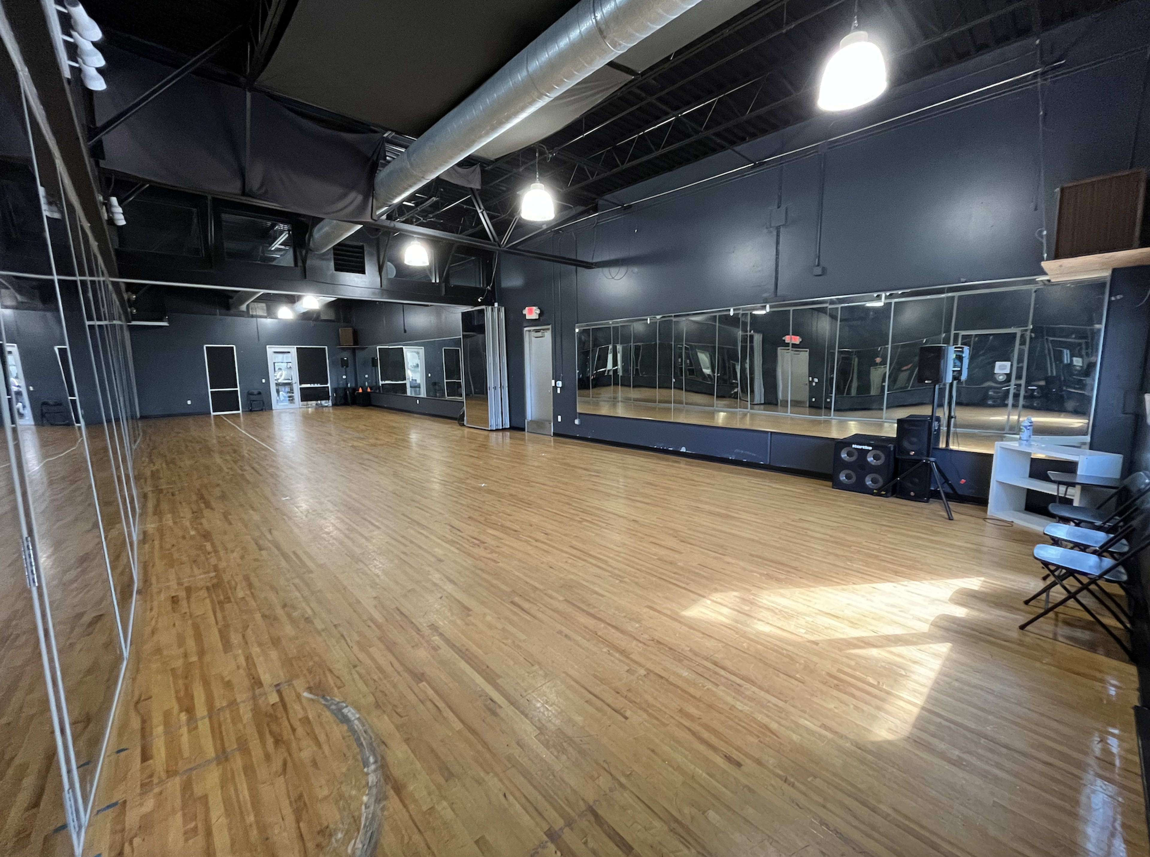 A spacious dance studio with wooden floors, large mirrors on one wall, and minimal furnishings including a speaker and folding chairs.