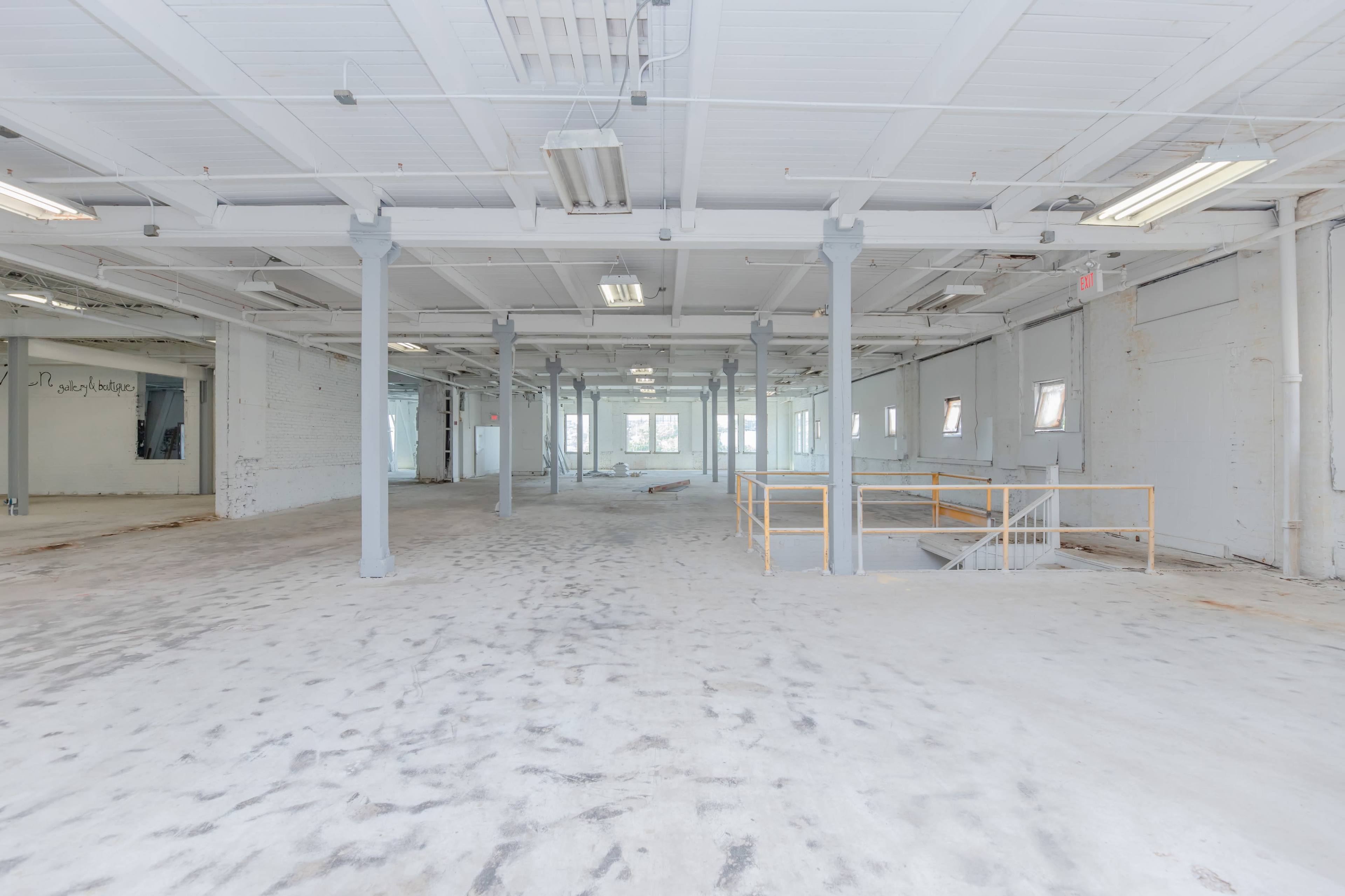 A spacious, empty industrial room with white walls, a concrete floor, and several windows along the sides.