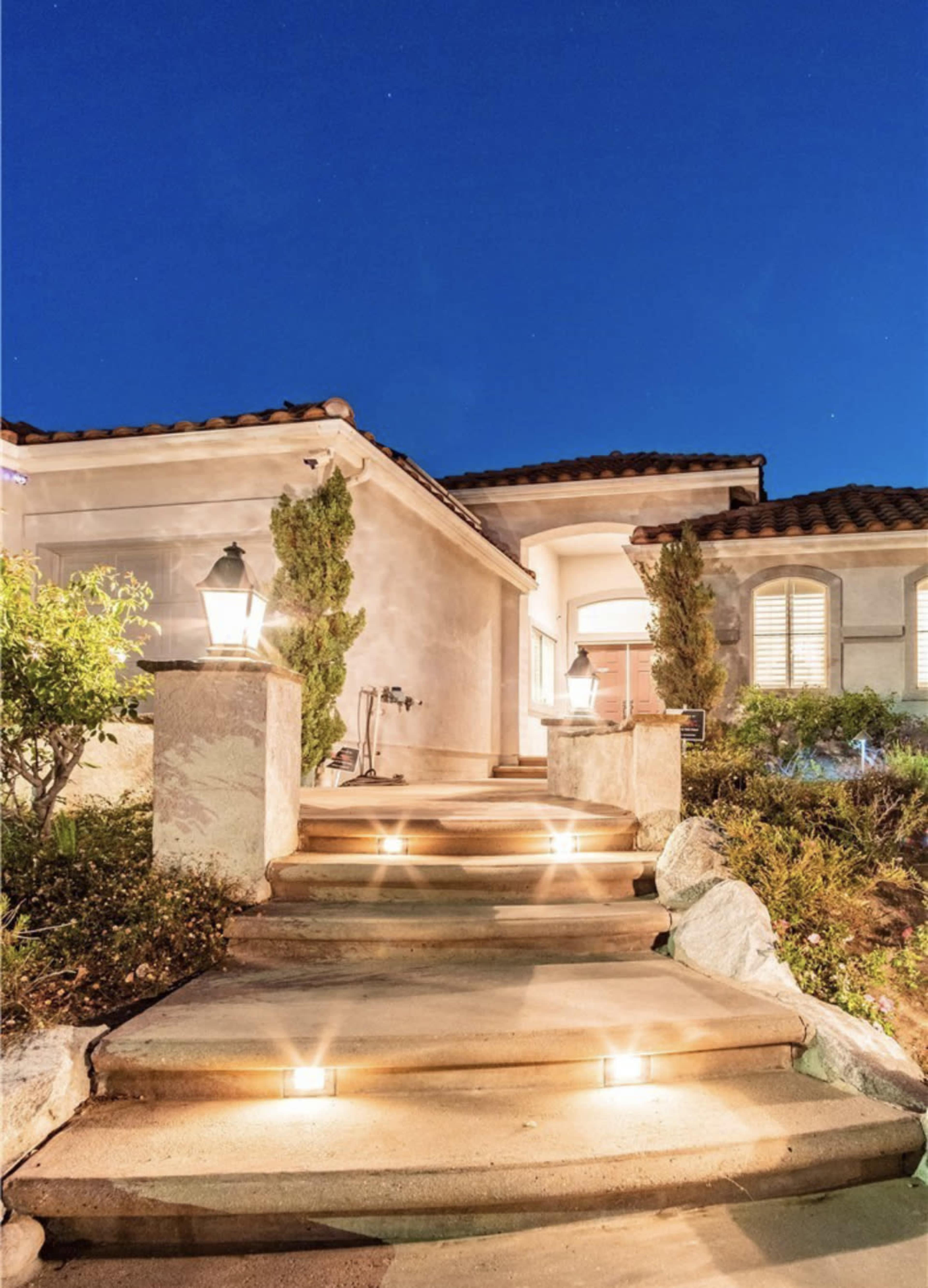 A well-lit pathway leads to the entrance of a house under a clear evening sky.