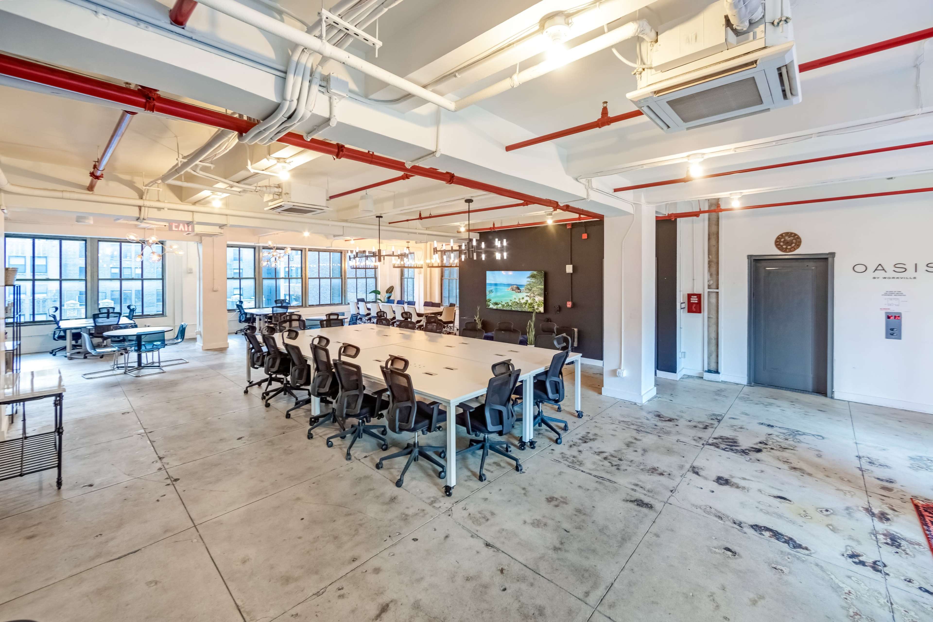 A modern office space features a long conference table surrounded by ergonomic chairs, with large windows providing natural light and a screen on the wall displaying a nature scene.