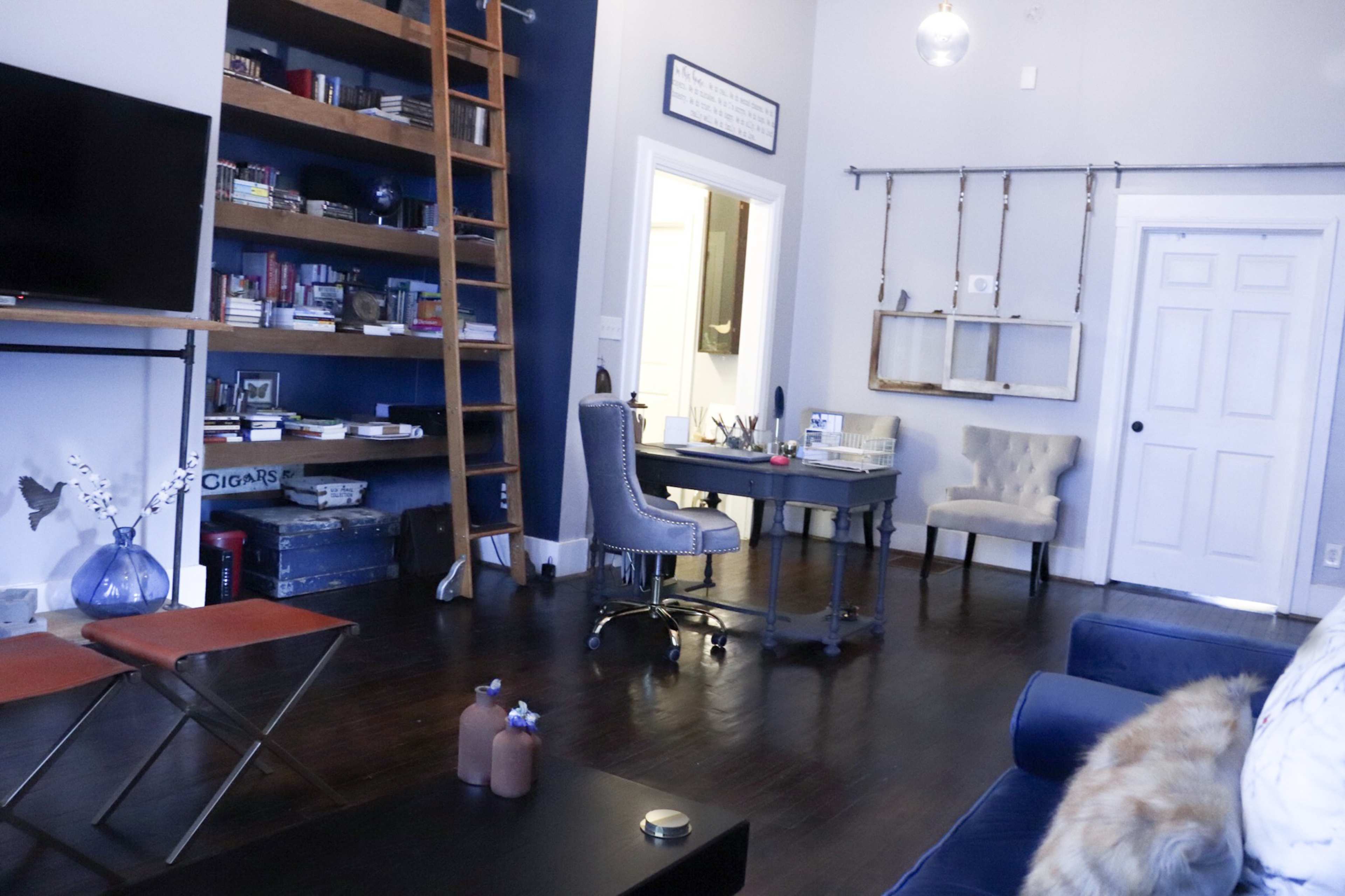 A modern living room with a blue couch, a desk with a chair, a wooden ladder leading to a bookshelf, and a door leading to another room.