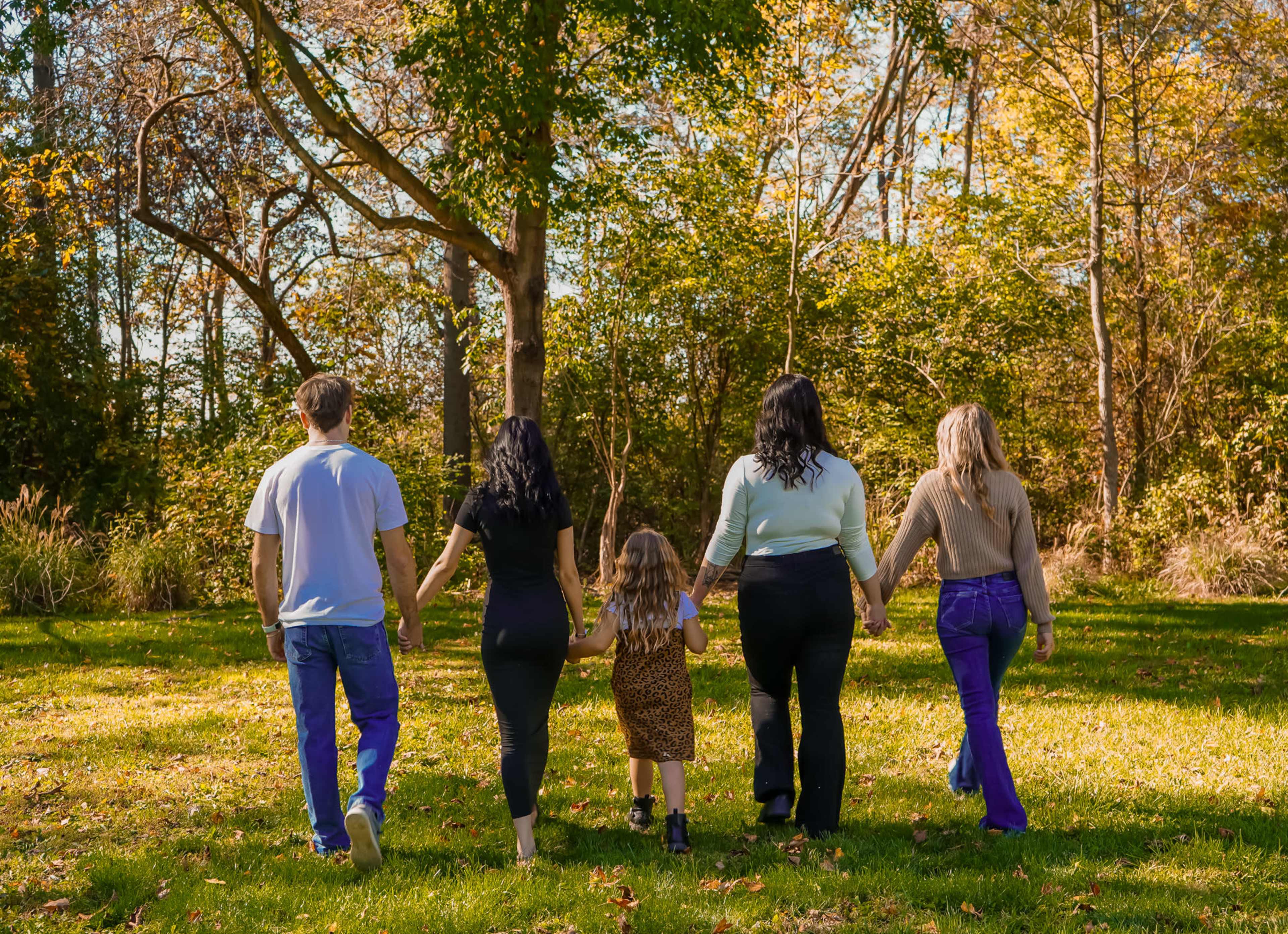 A group of five people walks hand-in-hand through a green park, surrounded by trees and autumn foliage.
