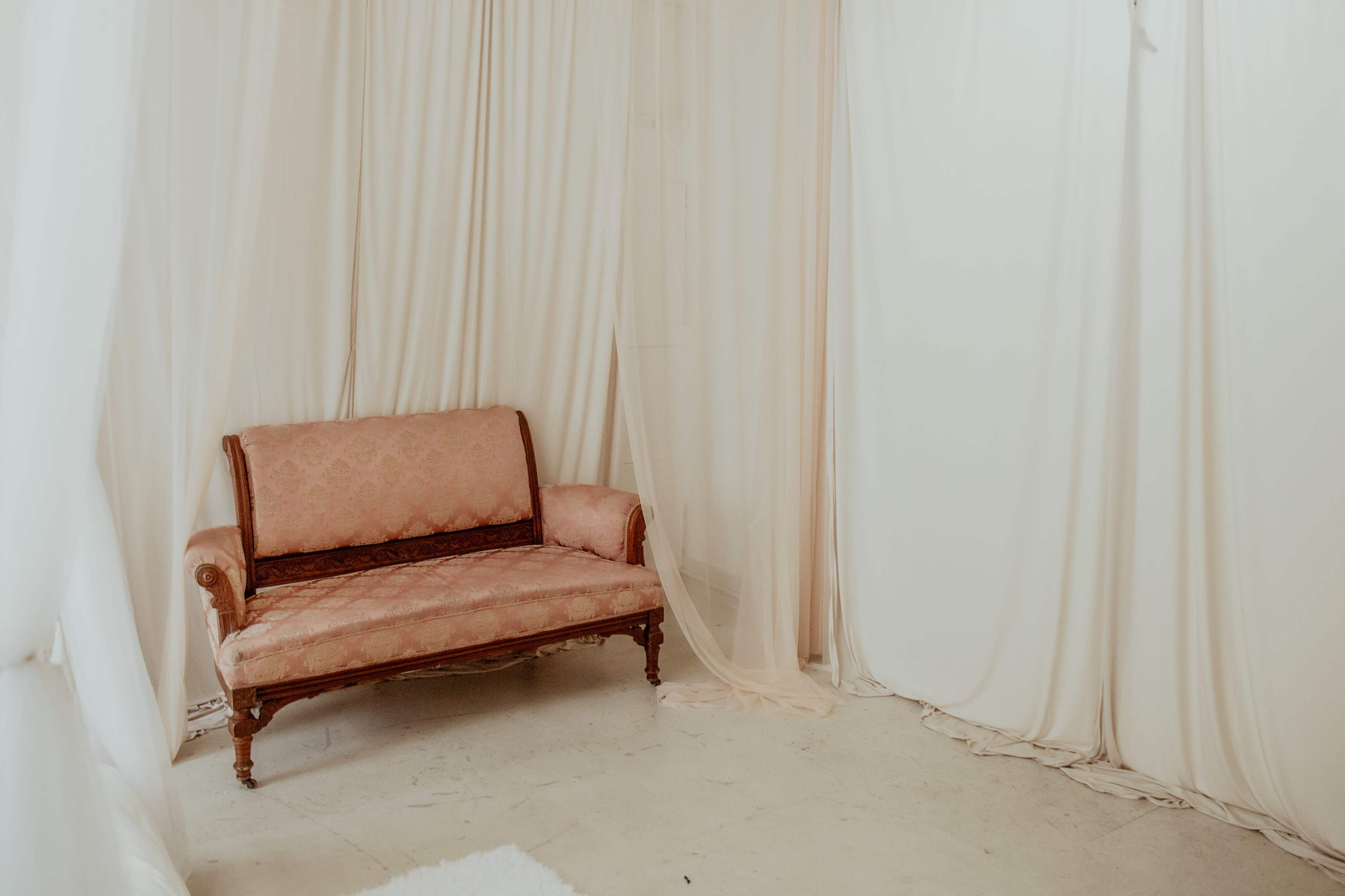 A pink upholstered loveseat sits near sheer, draped curtains in a softly lit room with a light-colored floor.