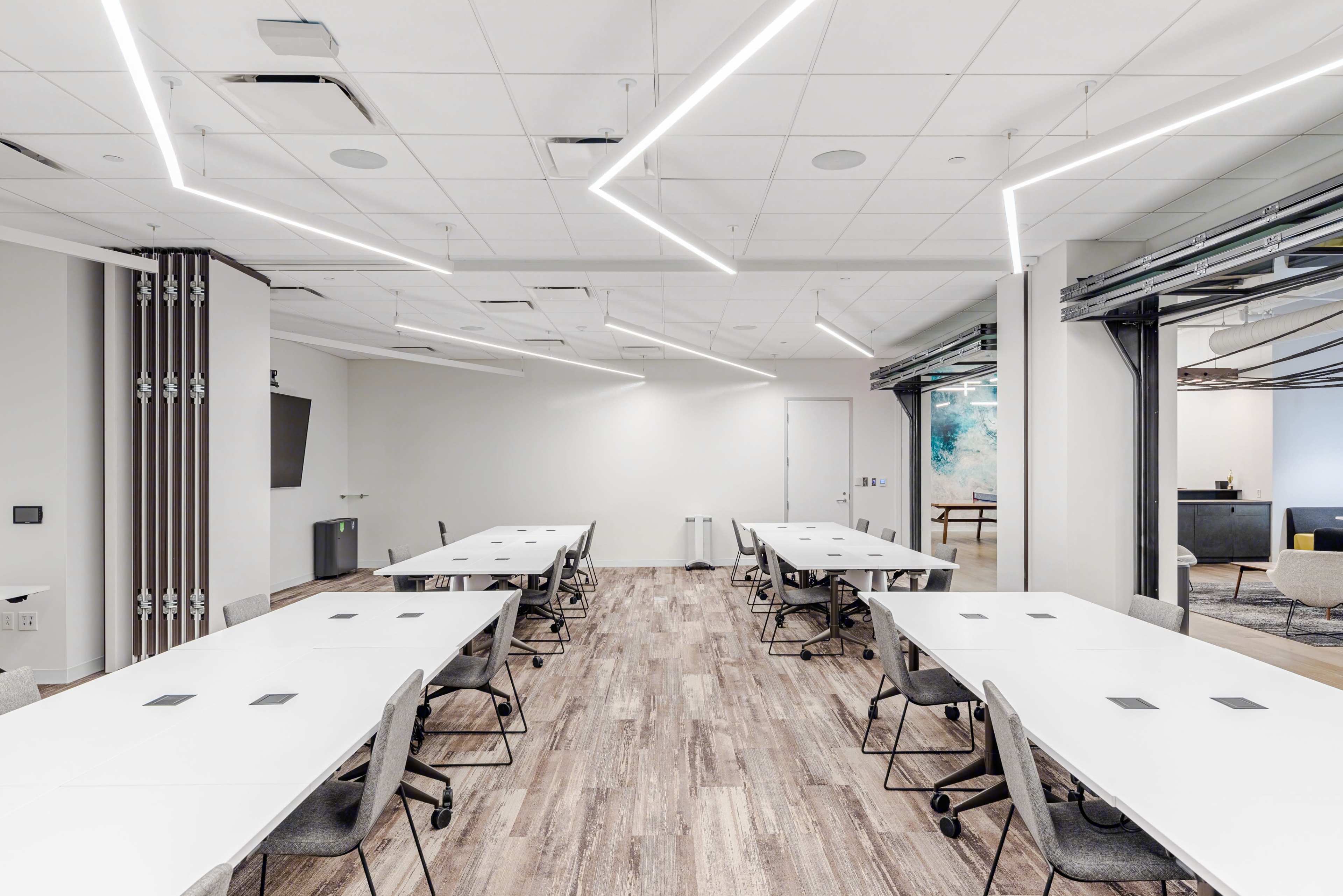 A modern conference room with multiple rectangular tables arranged in a collaborative layout, illuminated by angular ceiling lights.