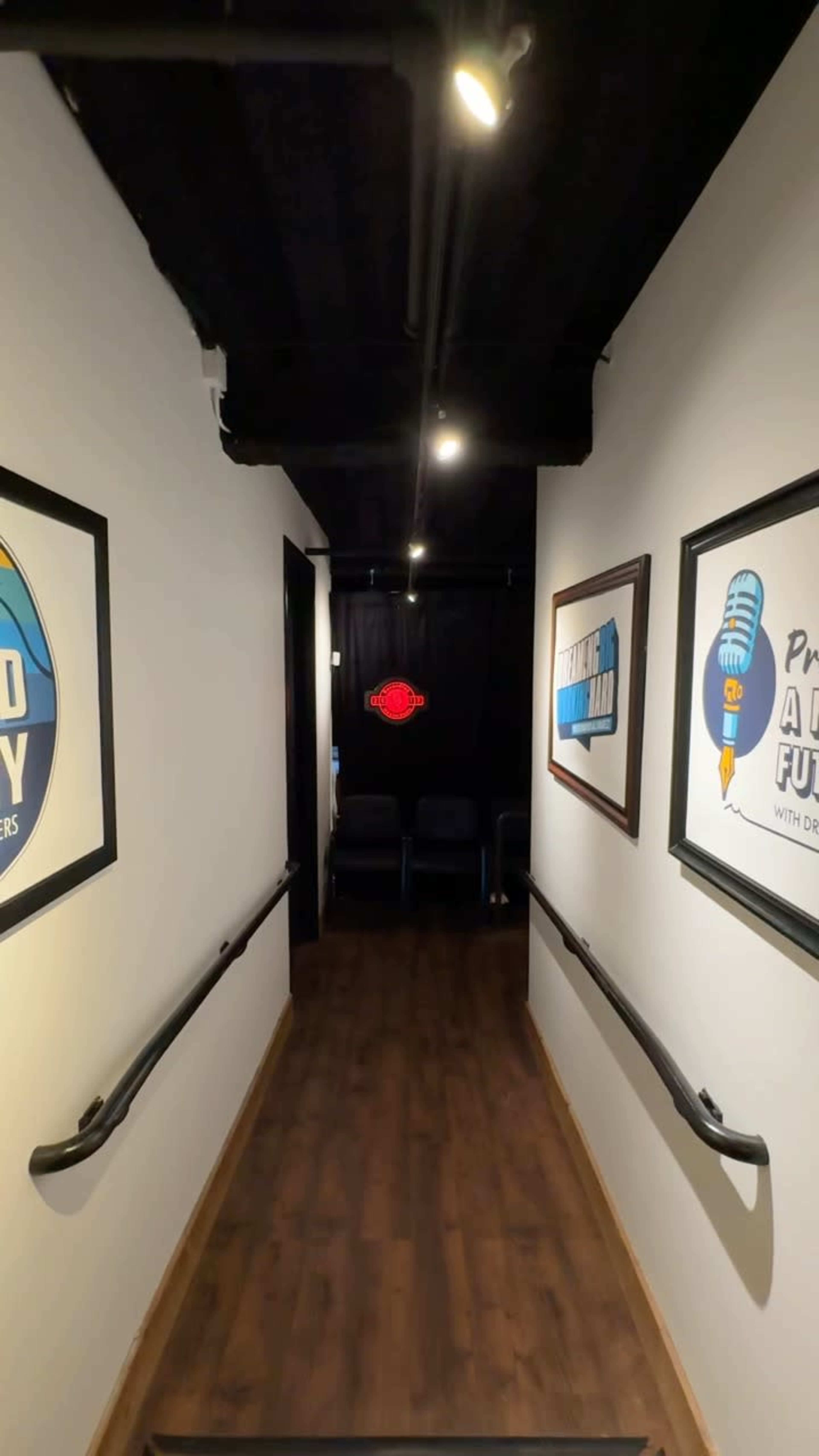 The image shows a long hallway with framed posters on the walls, leading to a doorway at the end with a red sign and a few chairs visible nearby.