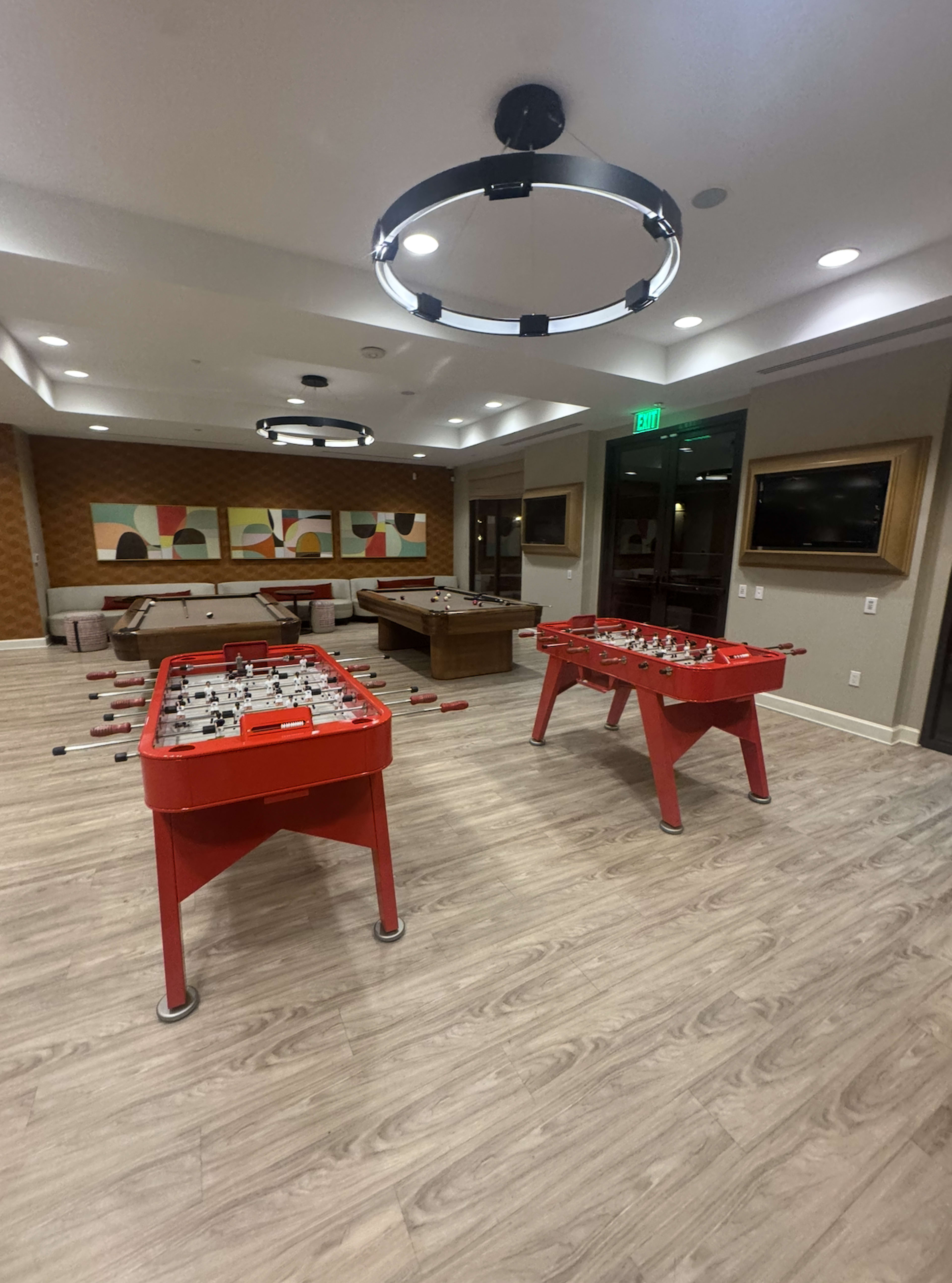The image shows a recreational room featuring two foosball tables and a billiards table, with modern lighting and decor.