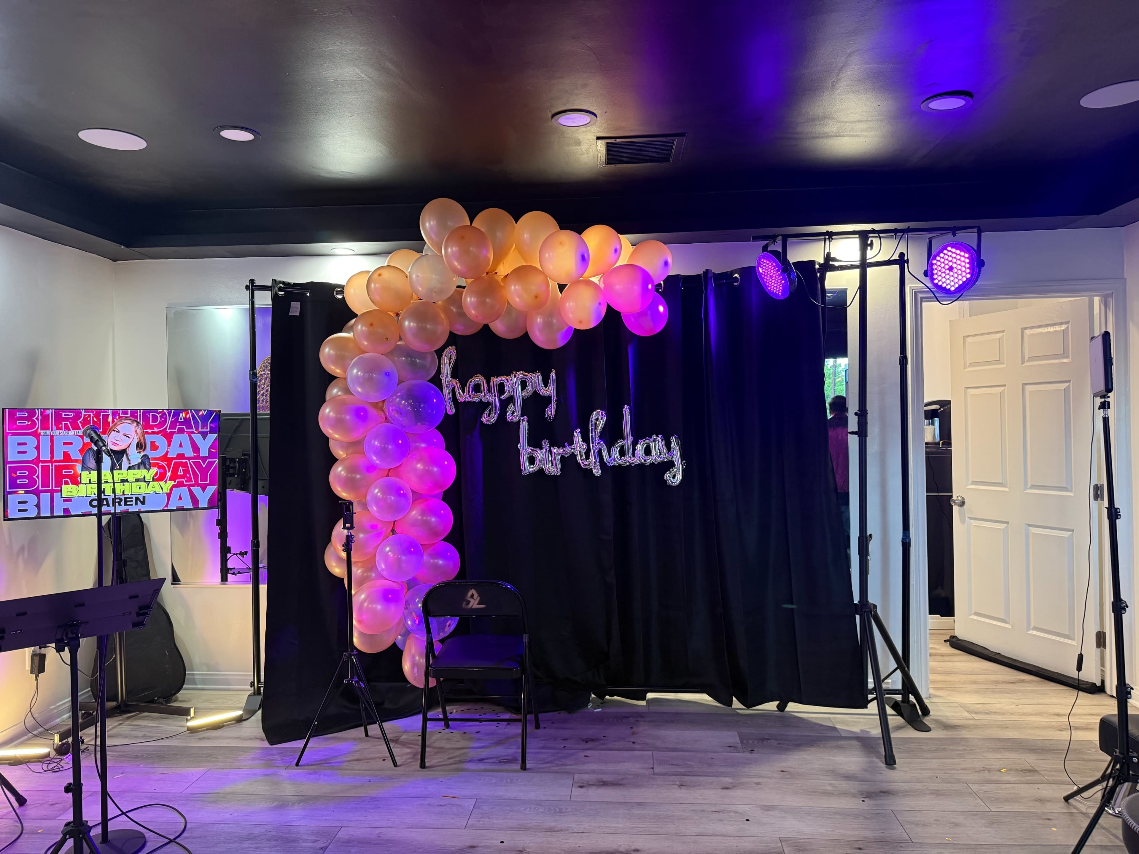 The image shows a decorated party space featuring a balloon arch and a "happy birthday" sign, with purple lighting and a stage setup in the background.