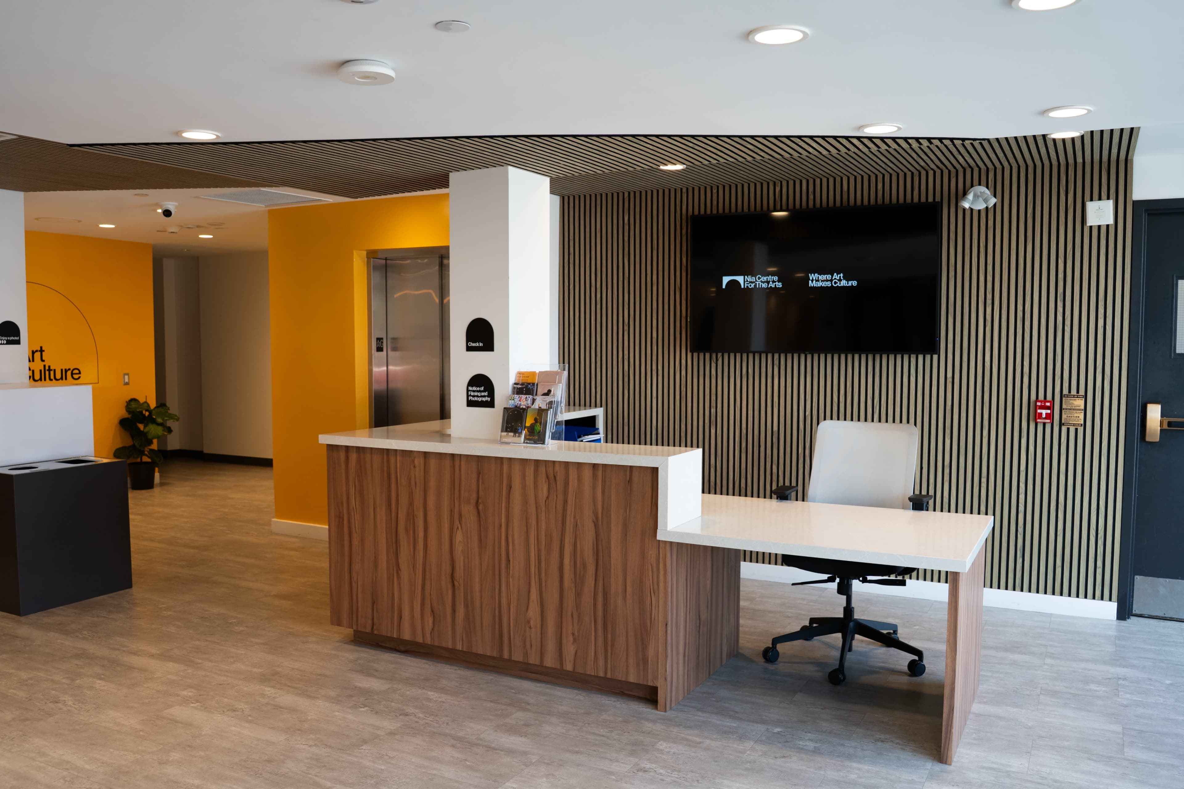 The image shows a modern reception area featuring a wooden desk, an elevator nearby, and a large screen on the wall displaying text.