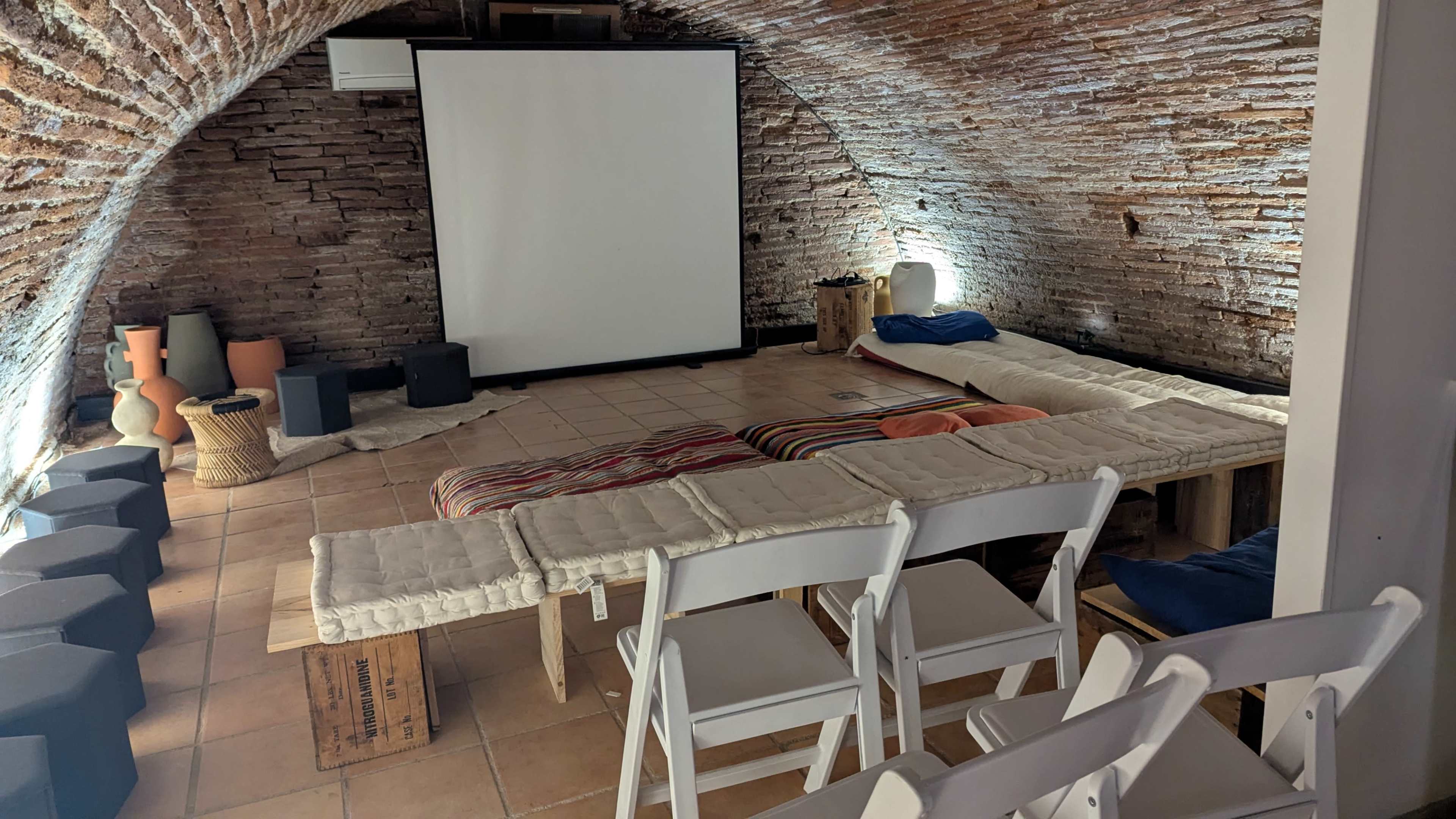 The image shows a cozy, arched room with a large screen at one end, several padded mats on the floor, and rows of white folding chairs arranged in front.